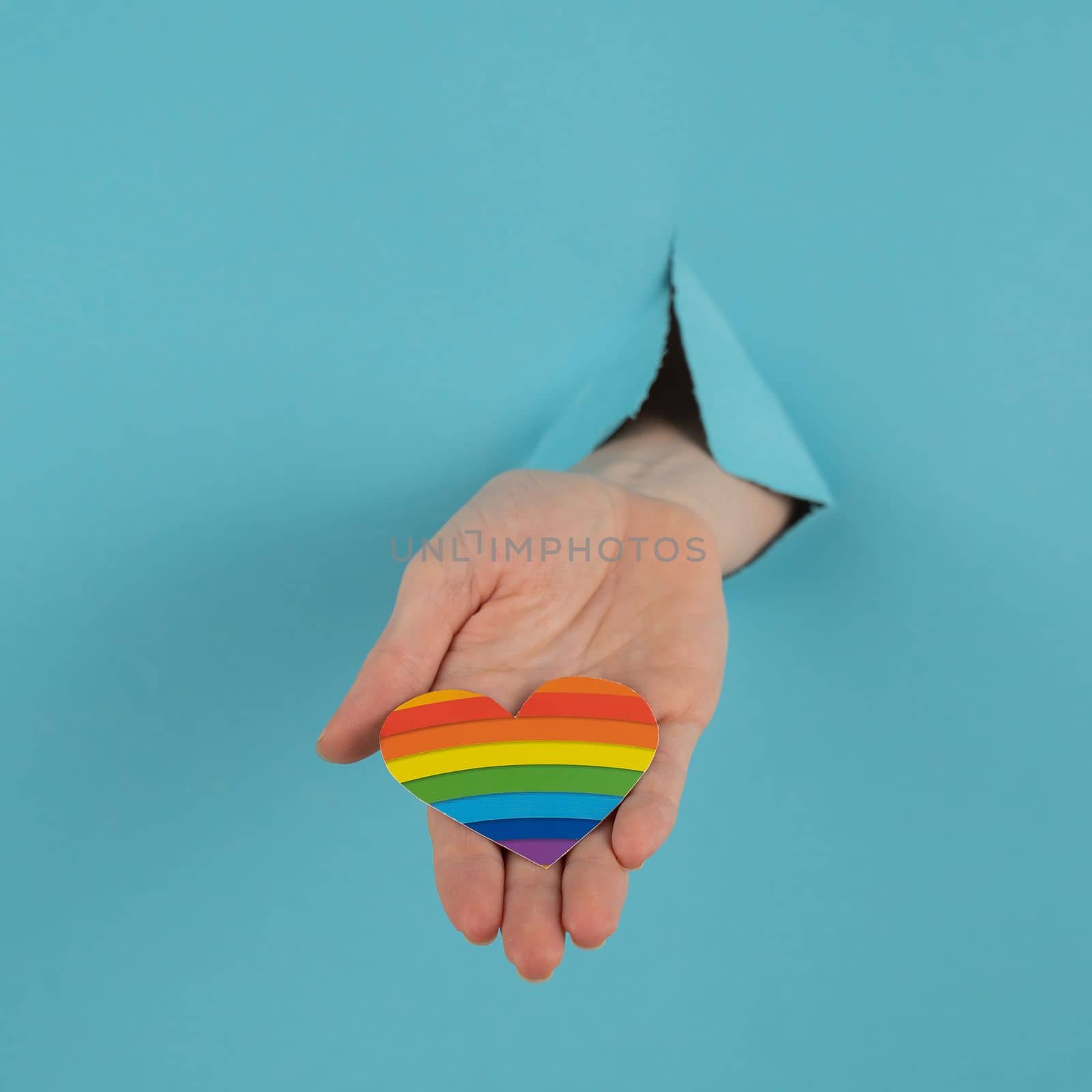 A hand with a rainbow-colored heart sticking out of a hole in a blue cardboard background