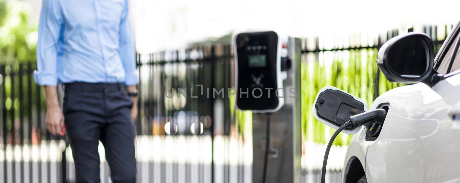 Focus closeup electric vehicle recharge battery at public charging station in the city area with blur businessman in background. EV car attached with electric charger for eco-friendly idea.