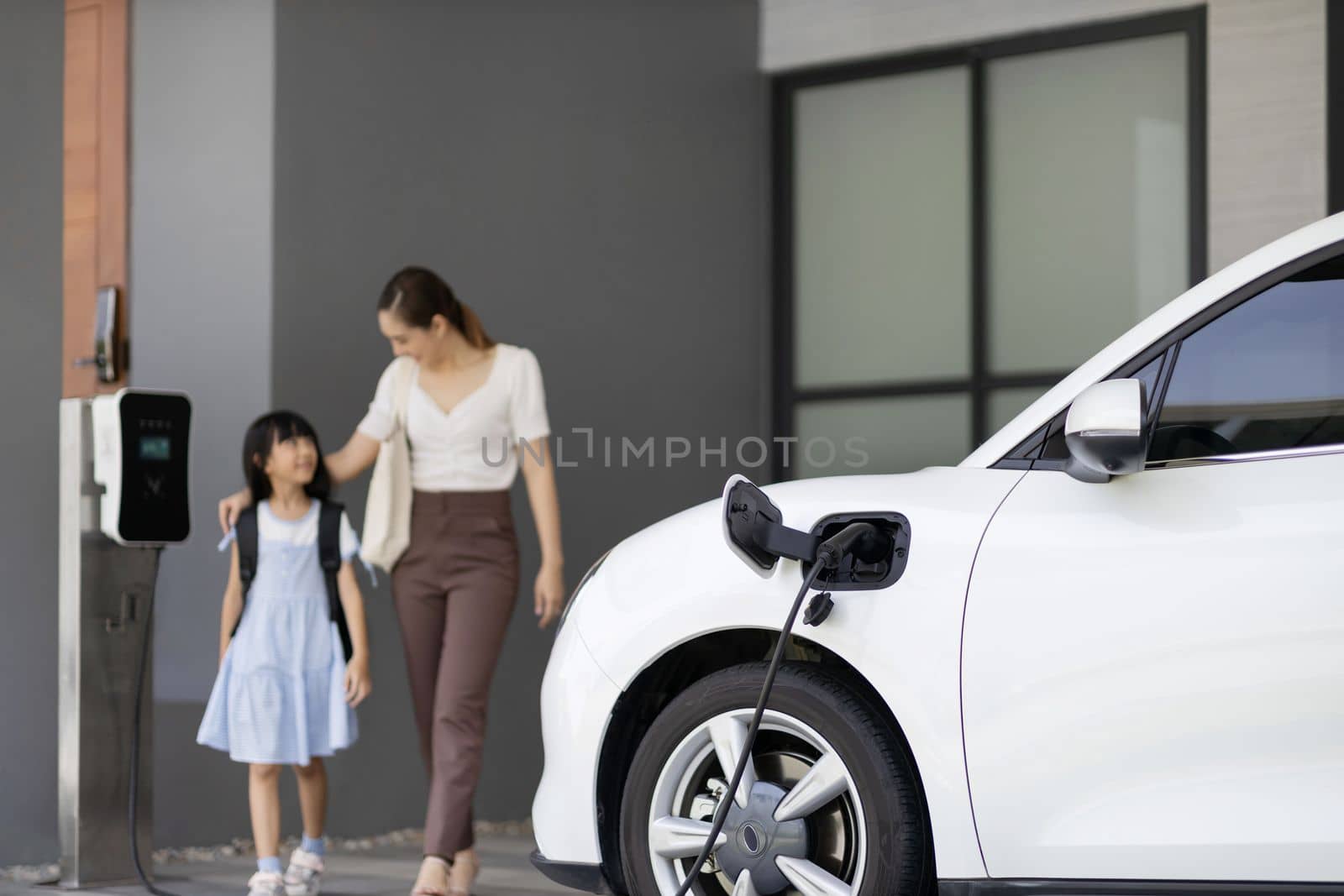Focus EV car recharging at home charging station with blurred progressive woman and young girl in background for alternative clean energy technology concept for renewable electric vehicle.