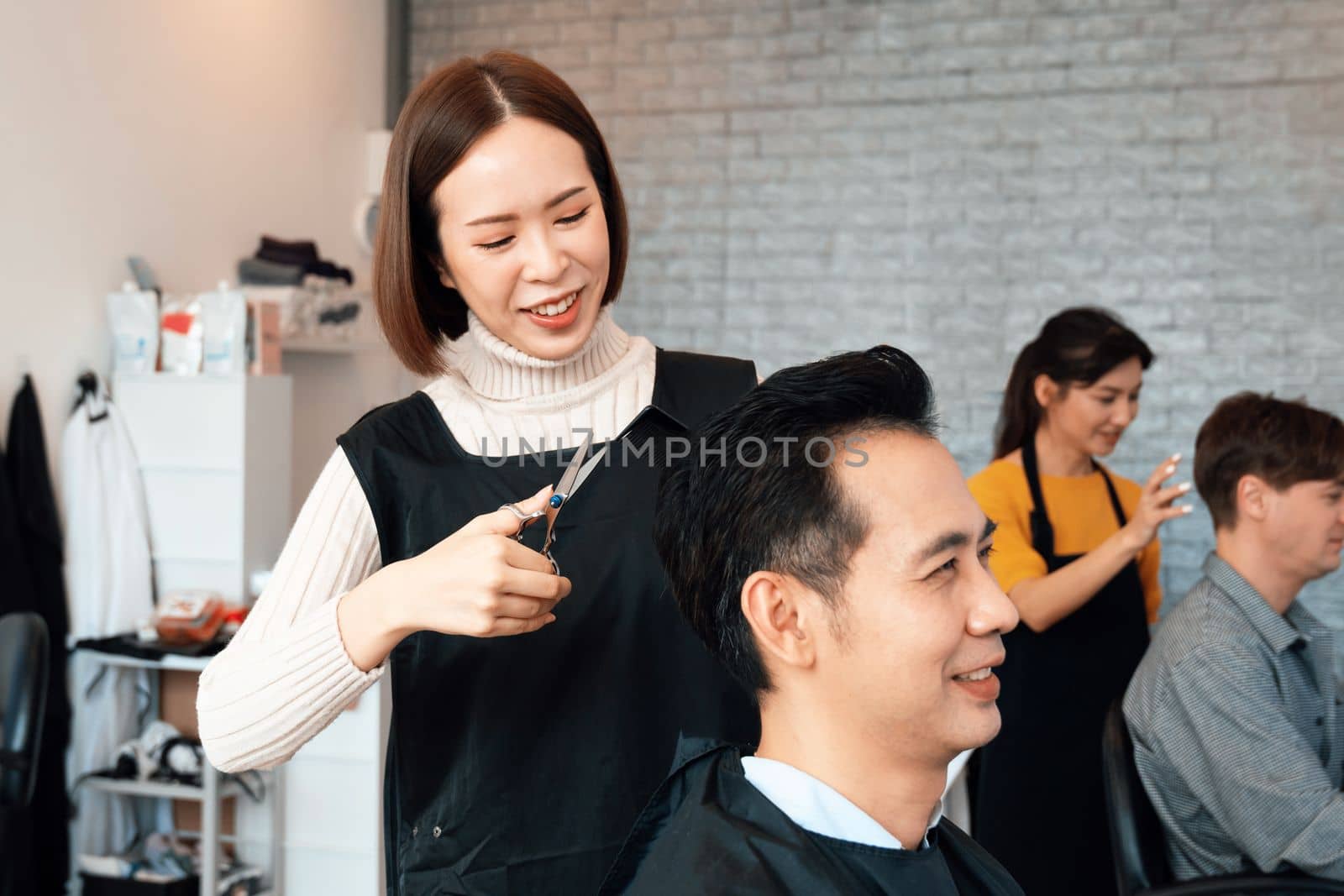 Asian female hairdresser making haircut for two male customer with male hairstyle in qualified barbershop. Men's hairstyling by a scissor and comb in hair salon concept.
