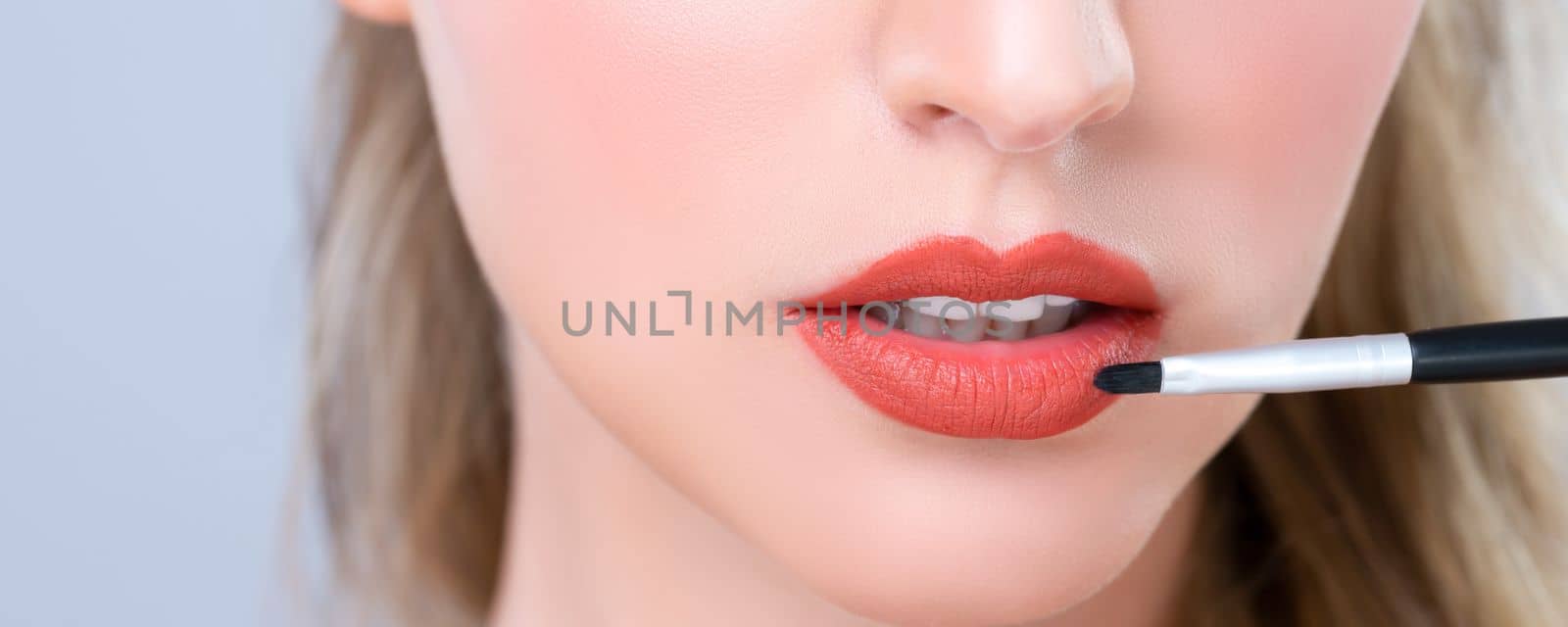 Closeup beautiful young woman with flawless healthy skin and natural makeup putting alluring fashion glossy red lipstick on her lip with lip brush in isolated background.