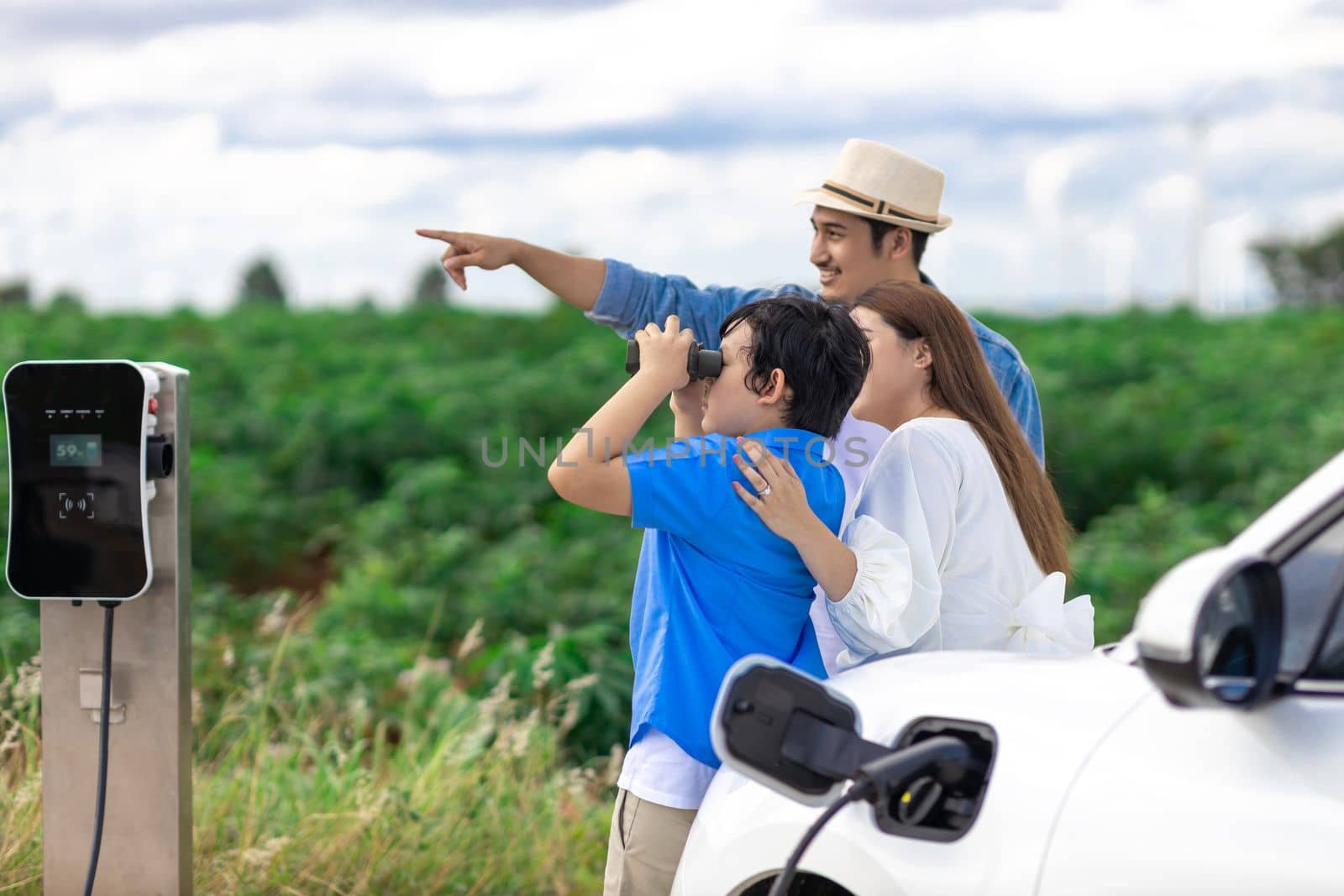 Concept of progressive happy family enjoying their time at wind farm with electric vehicle. Electric vehicle driven by clean renewable energy from wind turbine generator for charging station.
