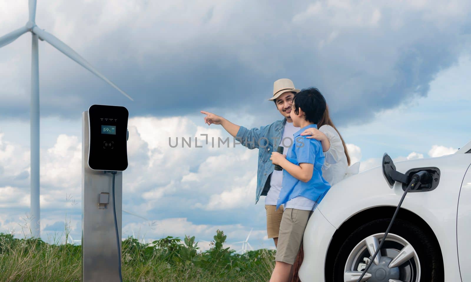 Concept of progressive happy family enjoying their time at wind farm with electric vehicle. Electric vehicle driven by clean renewable energy from wind turbine generator for charging station.