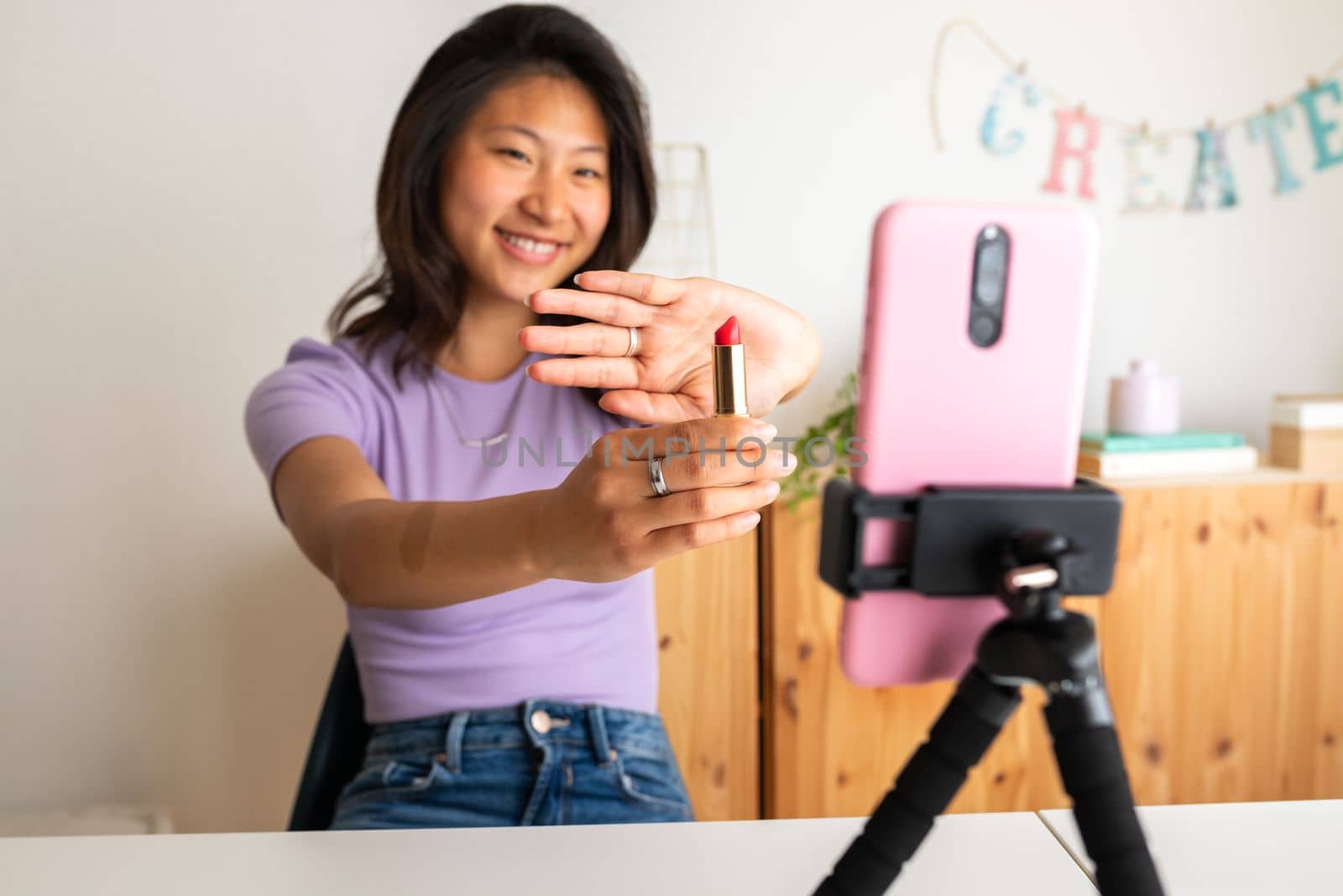 Happy young asian woman showing lipstick on camera, live streaming make up tutorial using phone on tripod. Social media and technology concepts.