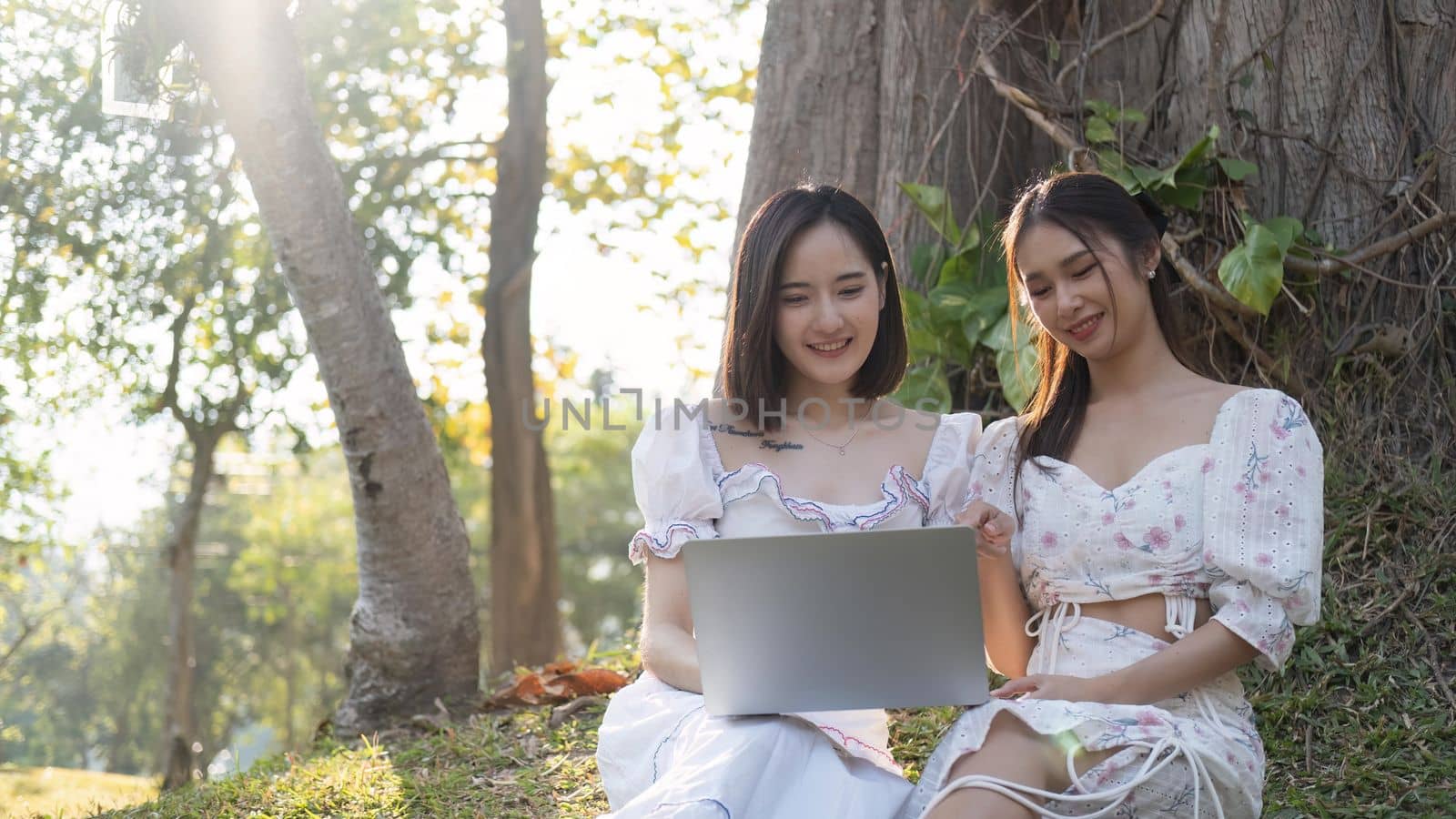 Two Asian women are watching a video on a laptop together while enjoying a picnic in the park by nateemee
