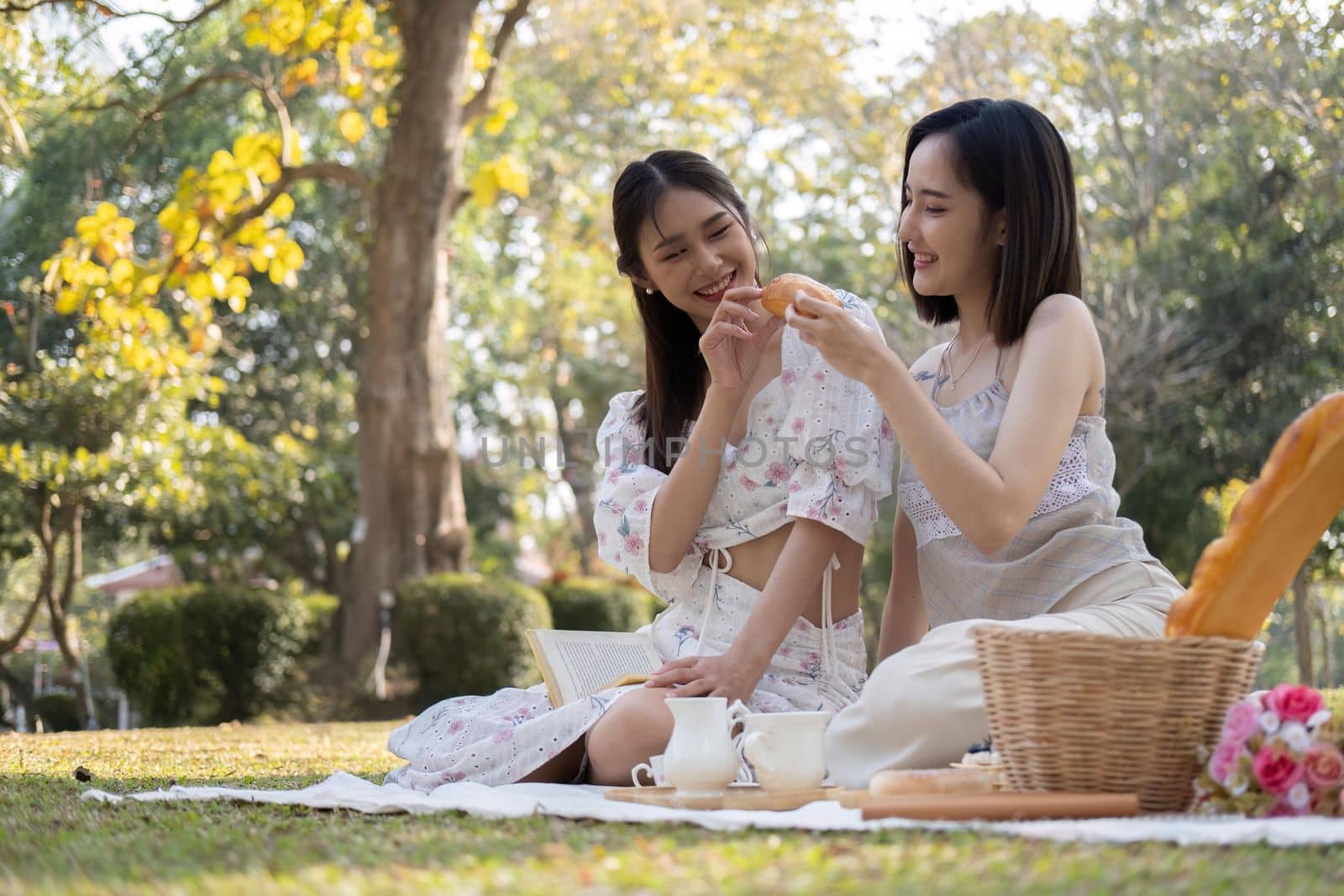 Two charming Asian women in beautiful dresses sitting on picnic cloth, having an tea picnic party together in the garden.
