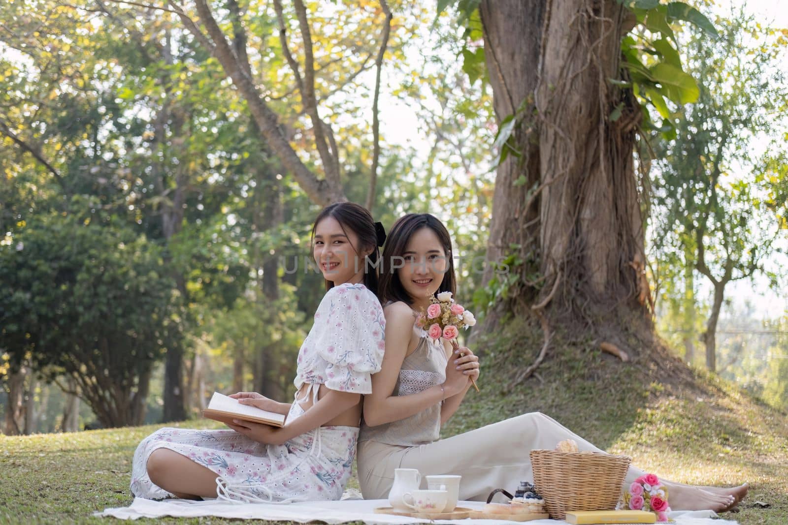 Charming beautiful young Asian woman in a lovely dress, holding a flower bouquet, enjoying a picnic with her friend in the park.