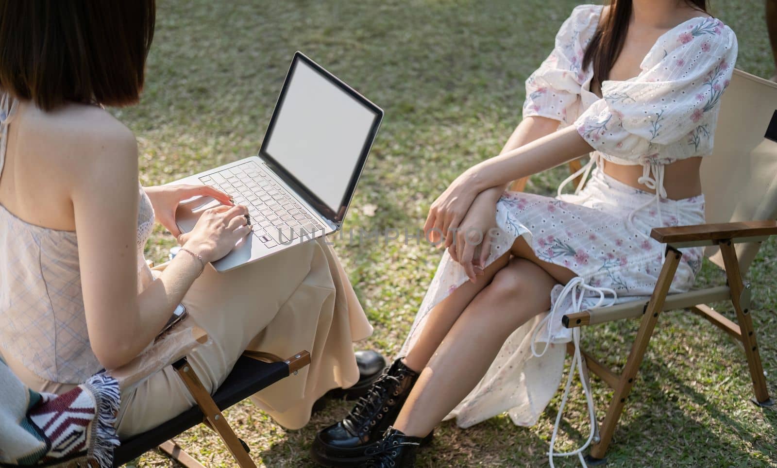 Two beautiful Asian women in lovely dresses enjoying picnic in the park, sipping , work on laptop sitting on picnic chairs by nateemee