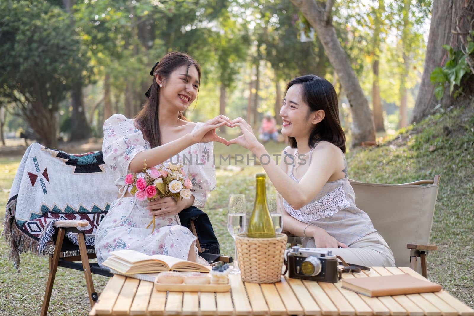 Asian woman friends, they are having picnic,they talk happily,charming Asian woman makes a heart hand sign with her friend while picnic in the park.