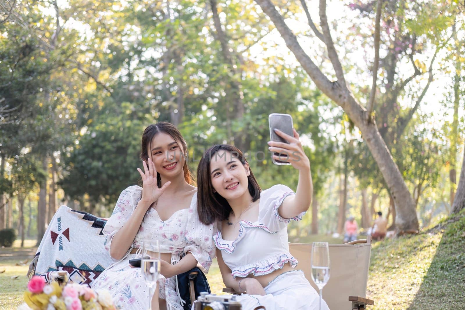 Two beautiful young Asian women picnic in the green park together, taking selfies with their phone they are having picnic,they talk happily by nateemee