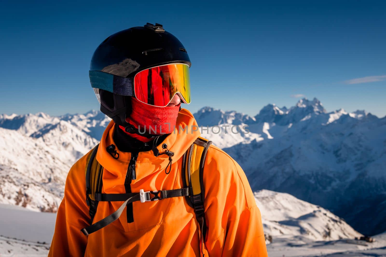 Portrait of a young man in a ski mask, stands in a ski resort against the backdrop of mountains and blue sky.