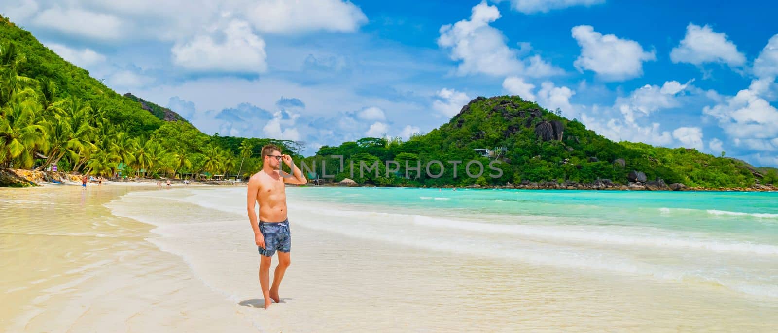 young tanning men in swim short at a white tropical beach with turquoise colored ocean Anse Volbert beach Praslin Seychelles.
