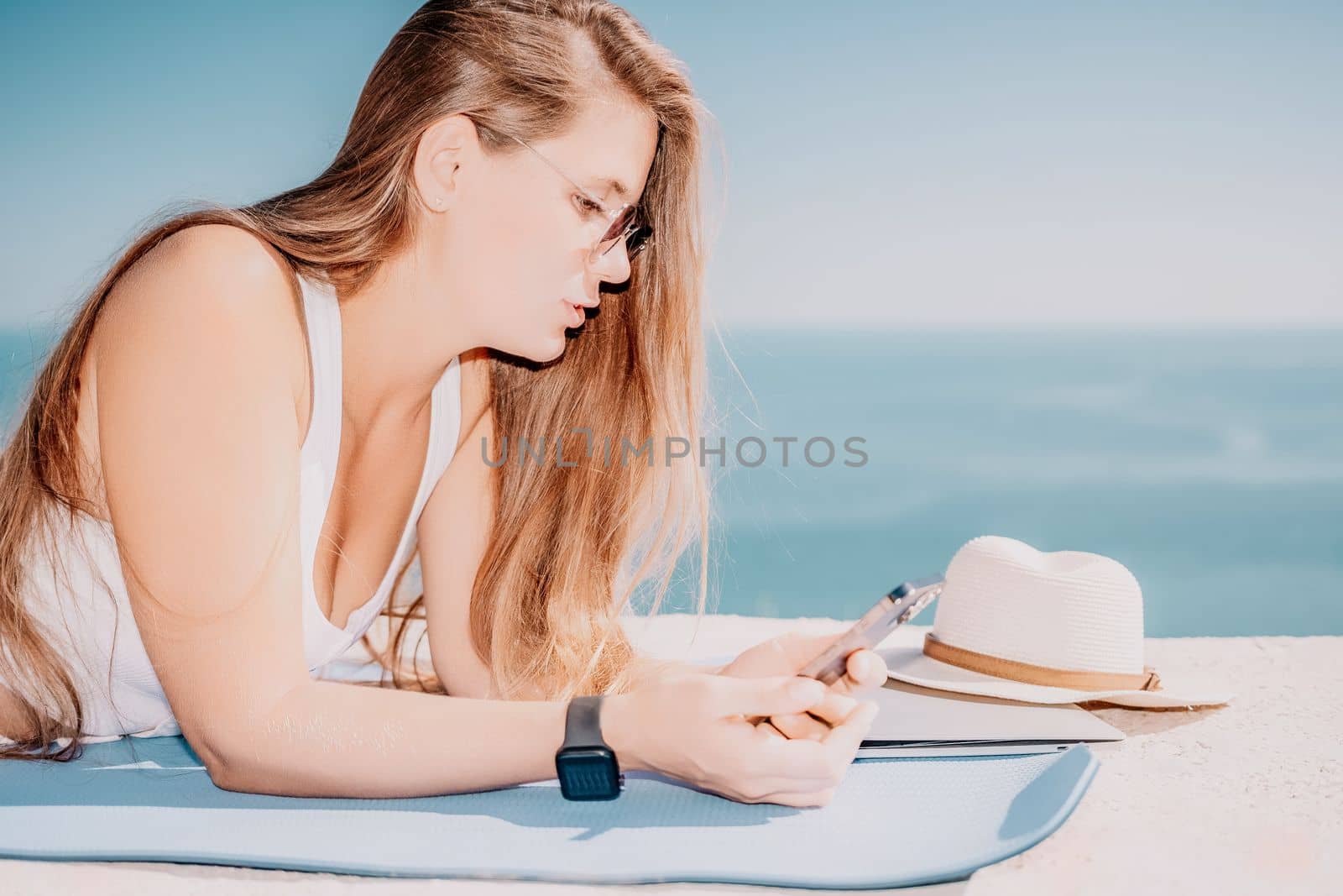 Successful business woman in yellow hat working on laptop by the sea. Pretty lady typing on computer at summer day outdoors. Freelance, travel and holidays concept.