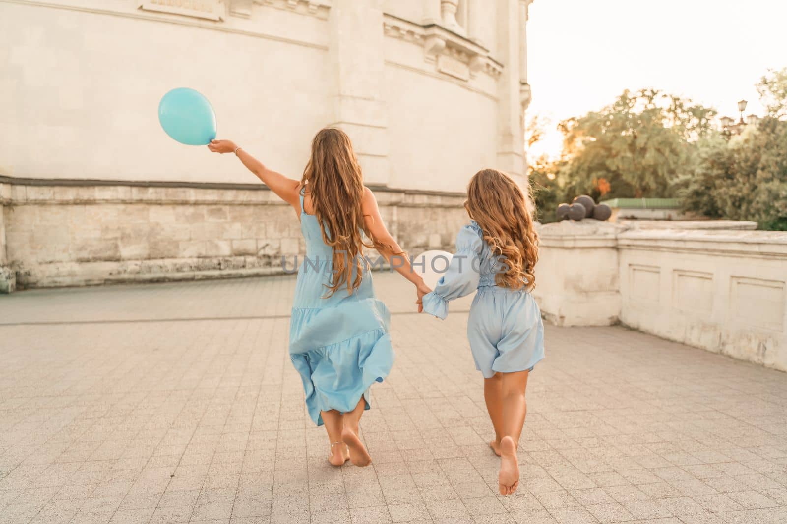 Daughter mother run holding hands. In blue dresses with flowing long hair, they hold balloons in their hands against the backdrop of a sunset and a white building. by Matiunina