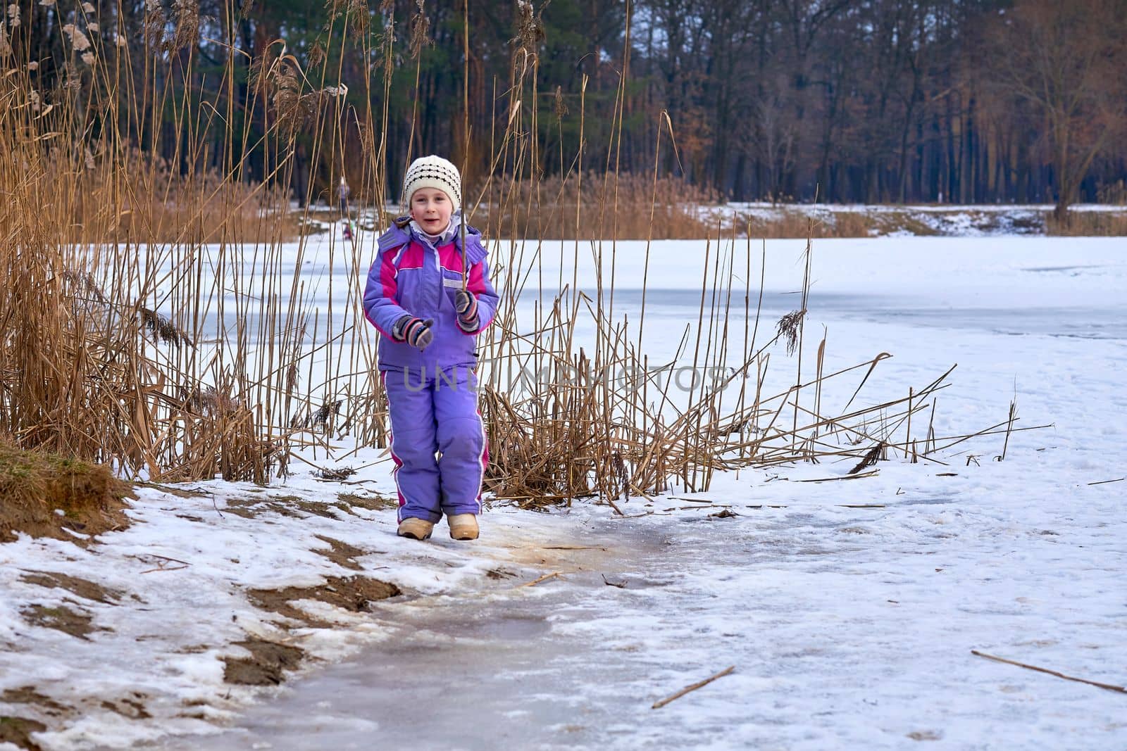 A cute child in a lilac jumpsuit walks on the lake in a winter park by jovani68