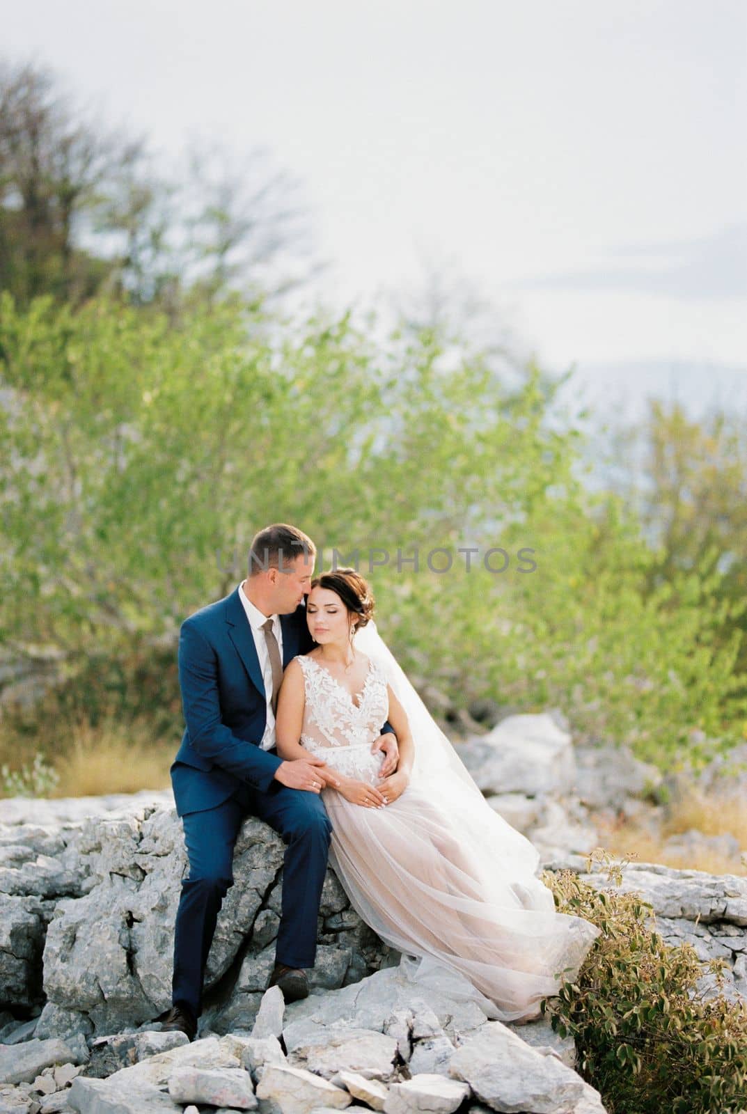 Groom hugs bride in a white dress sitting on the stones. High quality photo