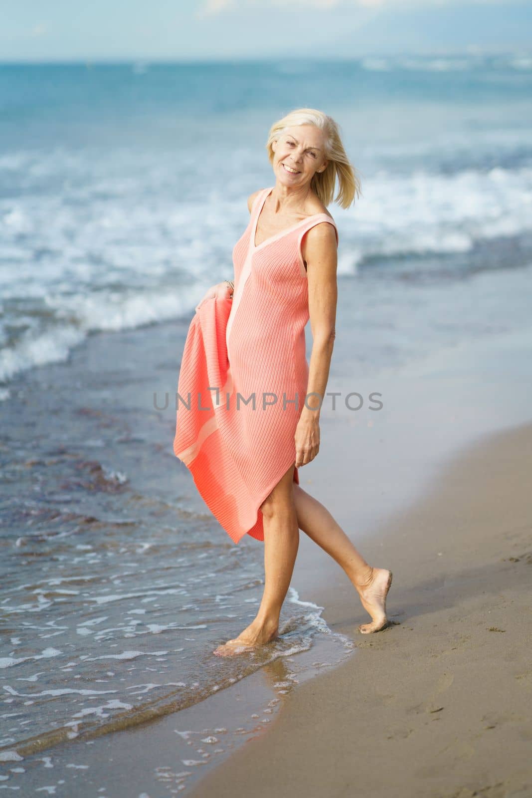 Beautiful mature woman walking along the shore of a tropical beach, wearing a nice orange dress. Elderly female enjoying her retirement at a seaside retreat.