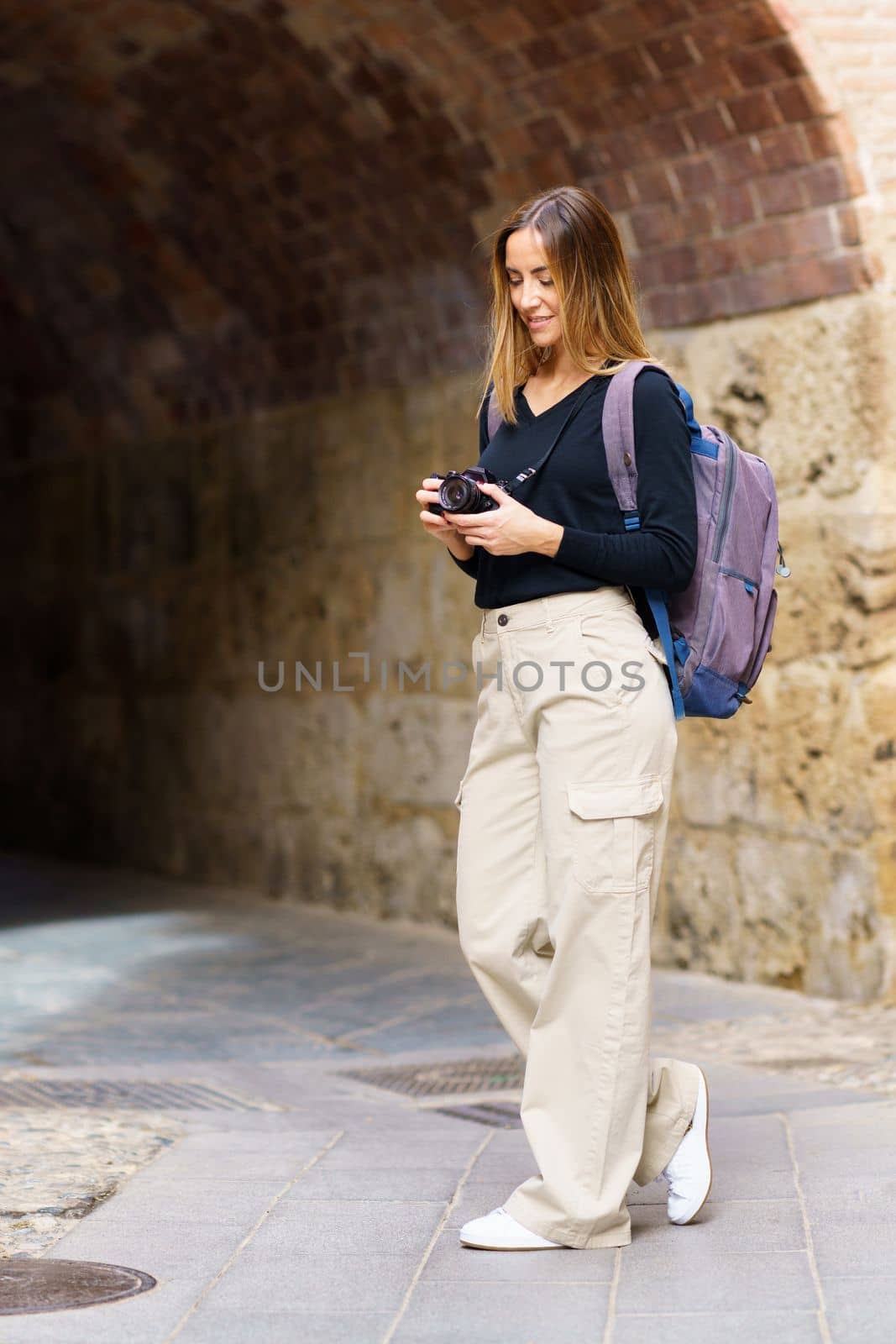 Side view of content young female traveler with long brown hair in casual clothes, and backpack taking photos on camera and smiling while standing near archway in old city during vacation