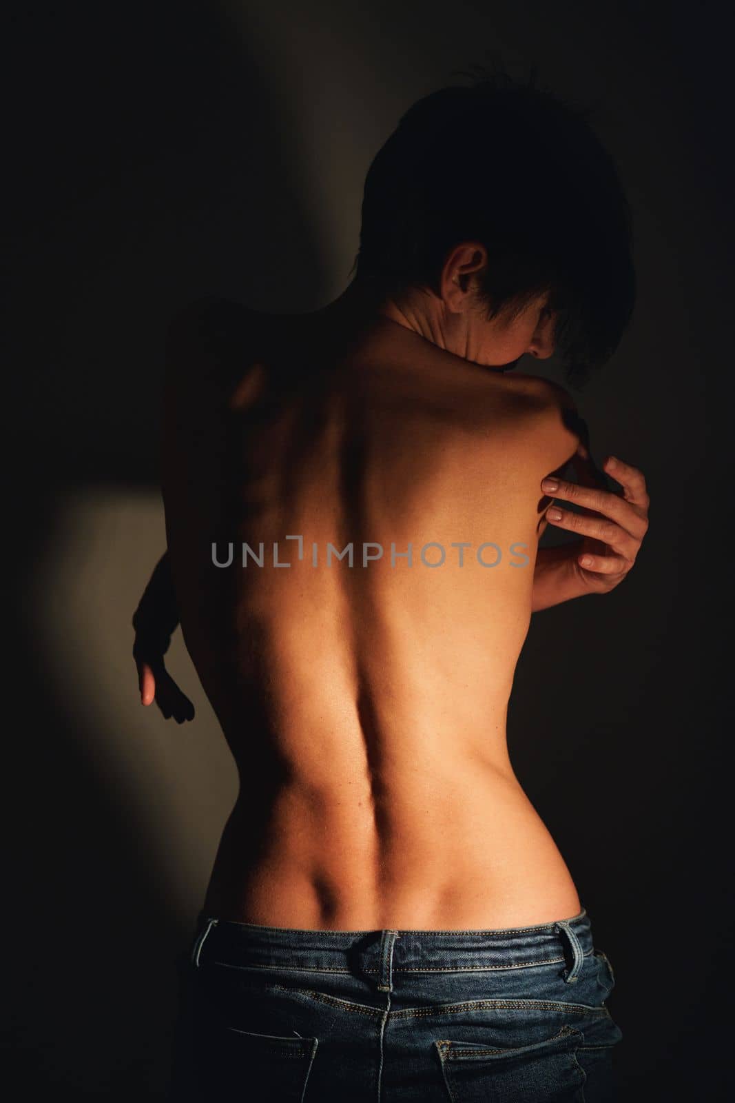 Back view of adult alluring topless woman with short dark hair in jeans standing near wall with crossed arms