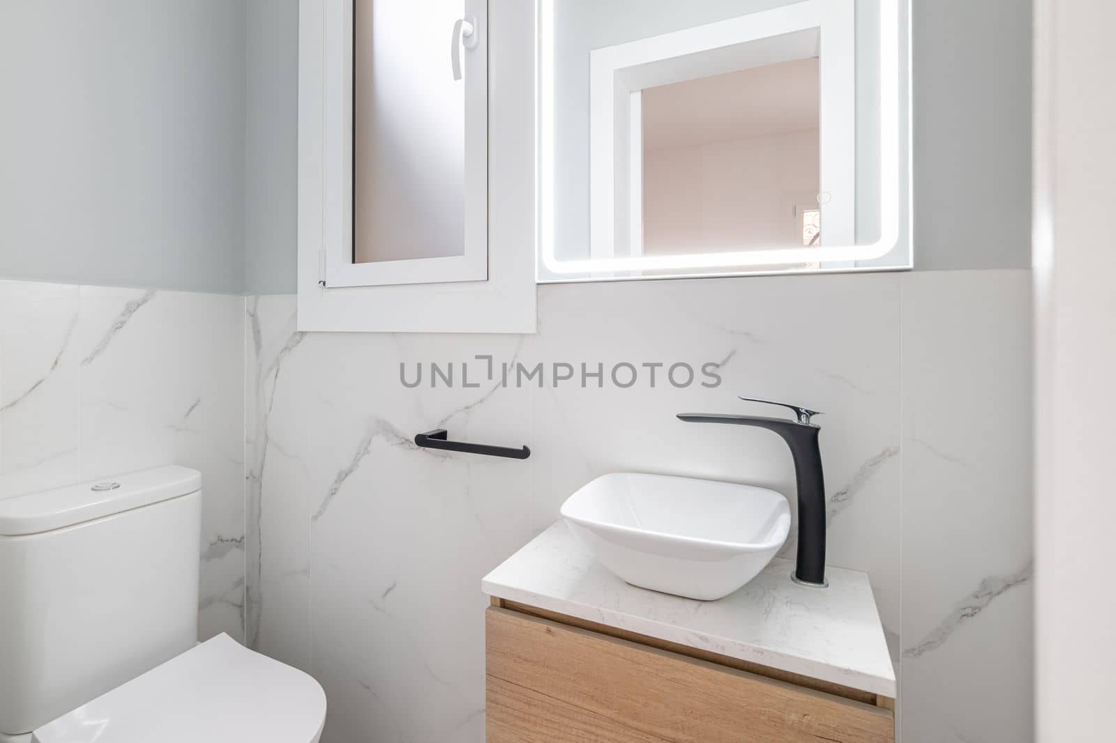 Small bathroom with toilet and designer sink on small vanity with black faucet. Walls of room are made of white natural marble tiles. Square mirror is illuminated with fluorescent light