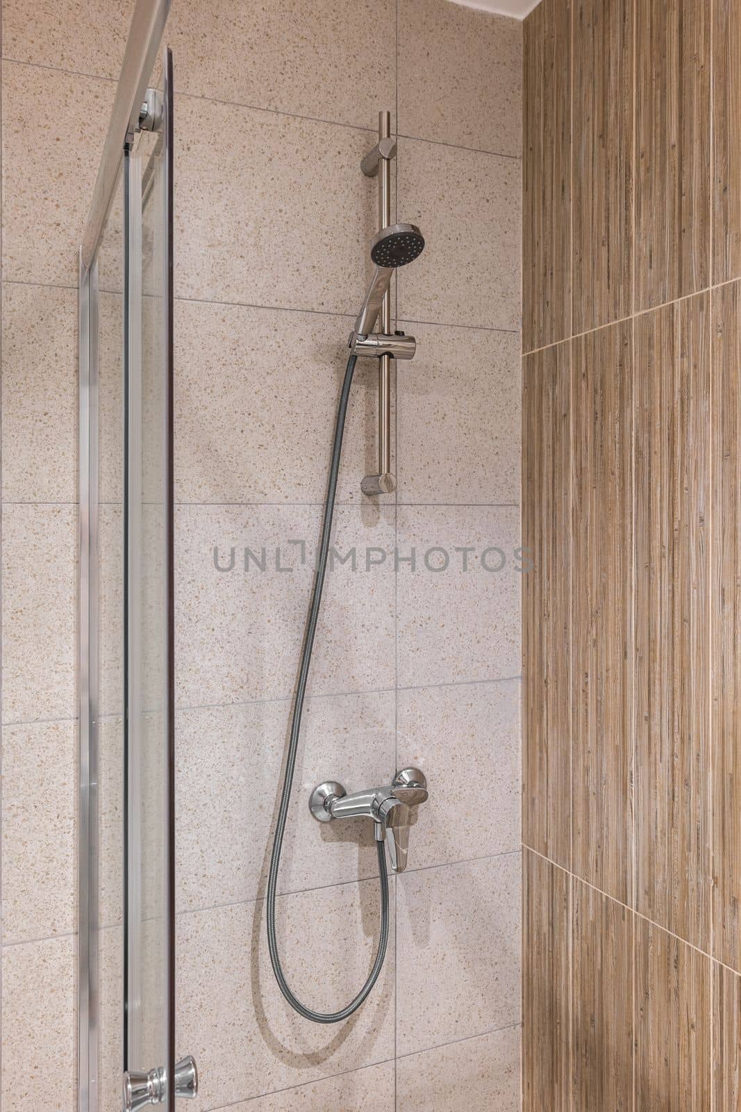 Closeup of a shower area with marble tiles on the walls. On the wall is a faucet with a long hose and a shower head. Area is fenced with glass doors to protect against splashing water. by apavlin