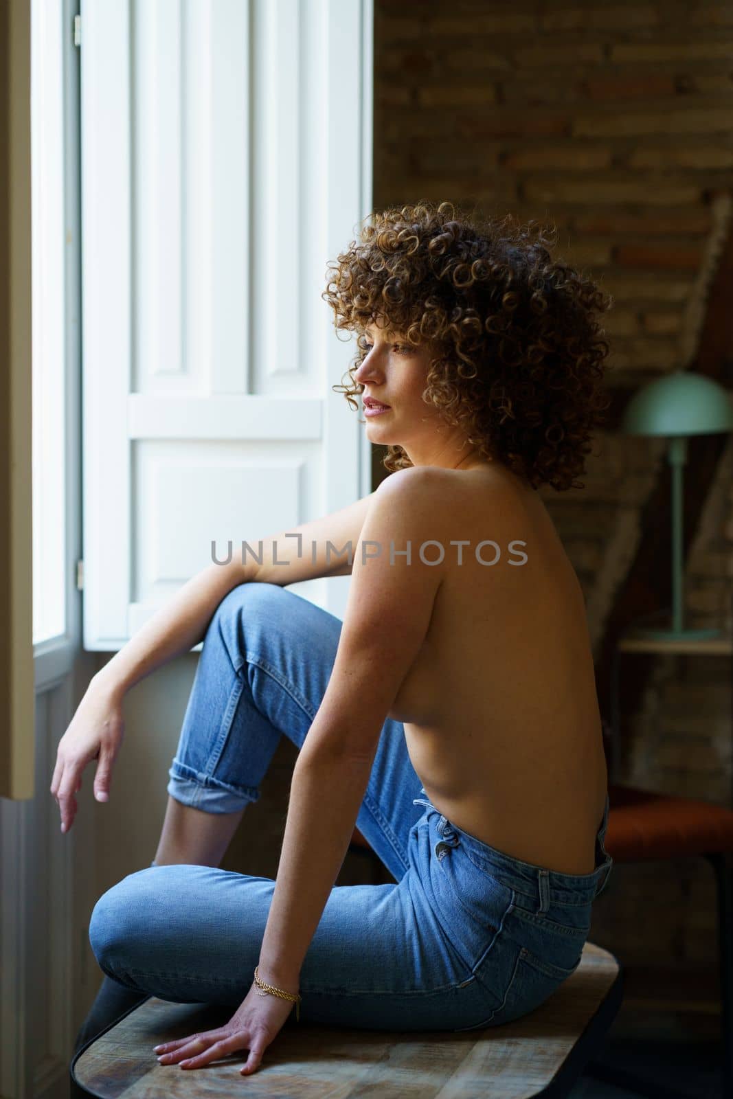 Side view of sensual young topless woman, with curly hair sitting on wooden stool with raised leg near window with shutters in room with brick wall