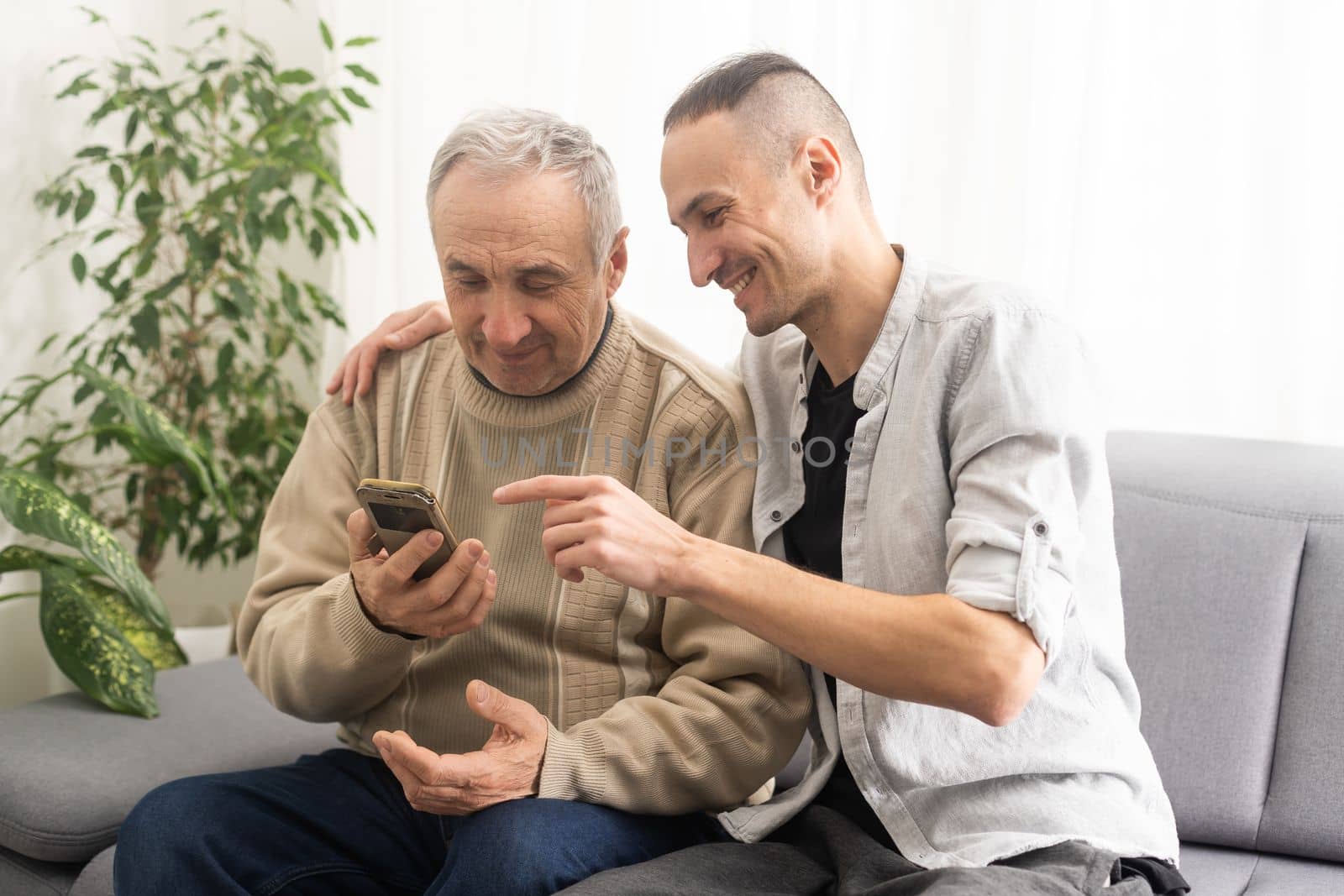 Happy two generations male family old senior mature father and smiling young adult grown son enjoying talking chatting bonding relaxing having friendly positive conversation sit on sofa at home