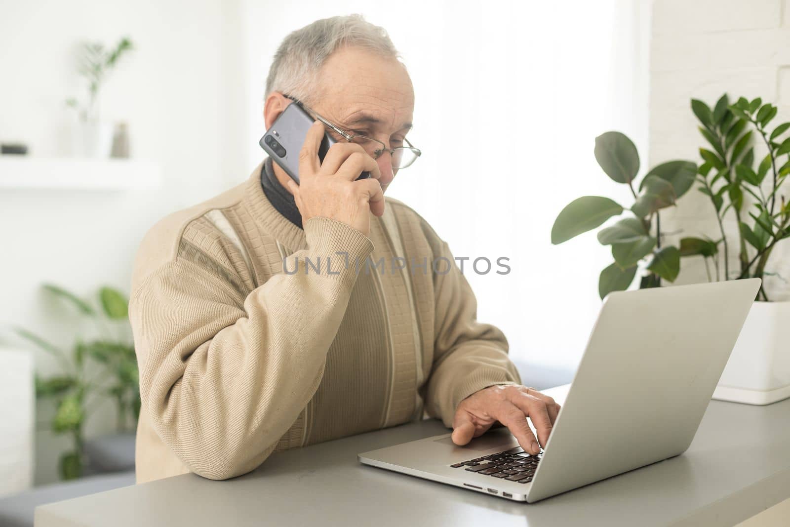 Handsome smiling senior man wearing glasses using mobile phone while sitting at his cozy workplace with laptop at home, retired male chatting with friends in social media, typing on smartphone