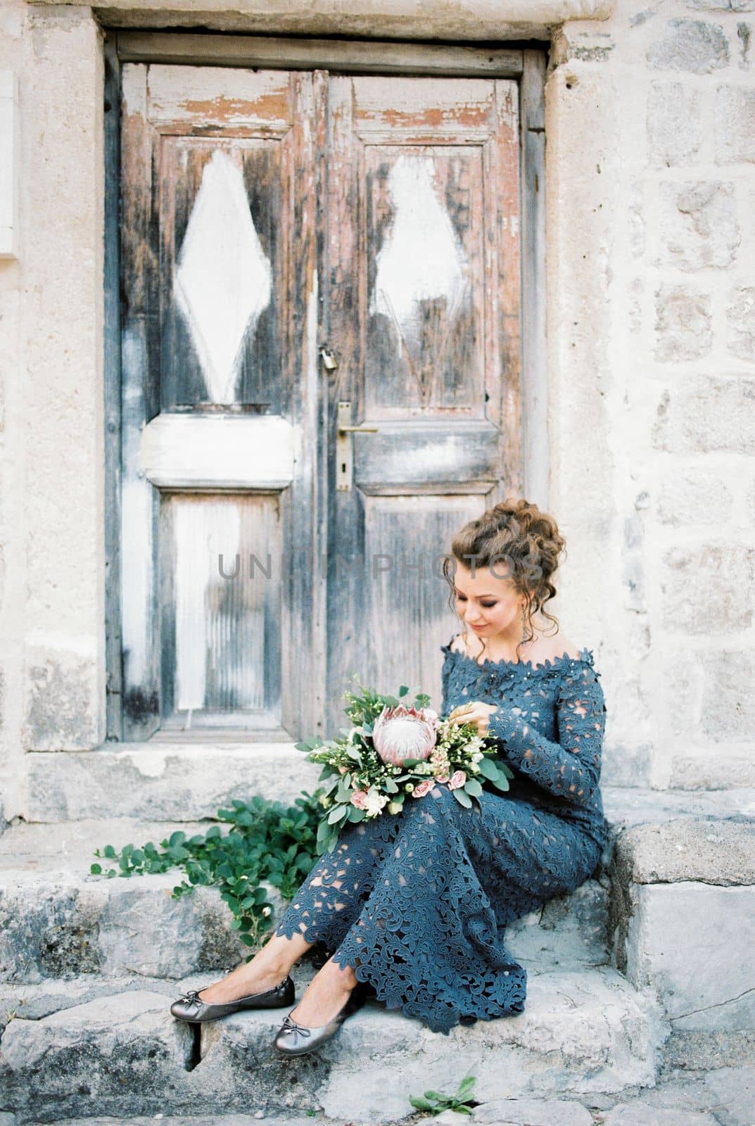 Bride with a bouquet of flowers on her knees sits on the steps of an old building near the door. High quality photo