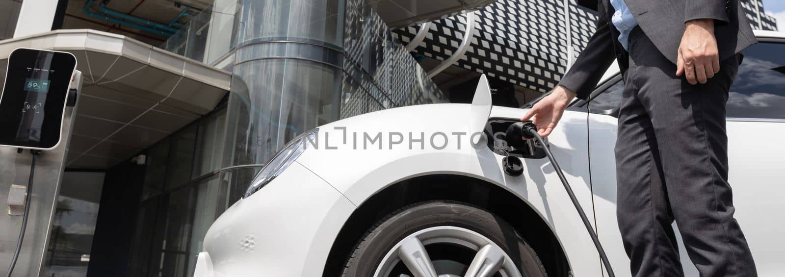 Closeup progressive man holding EV charger plug from public charging station for electric vehicle with background of residential building as concept eco-friendly sustainability energy car concept.