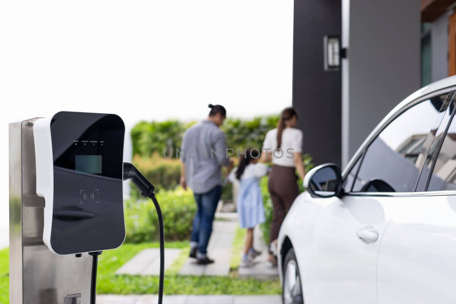 Focus closeup electric vehicle recharging battery from home electric charging station with blurred family in background. Renewable clean energy car for progressive eco awareness lifestyle concept.