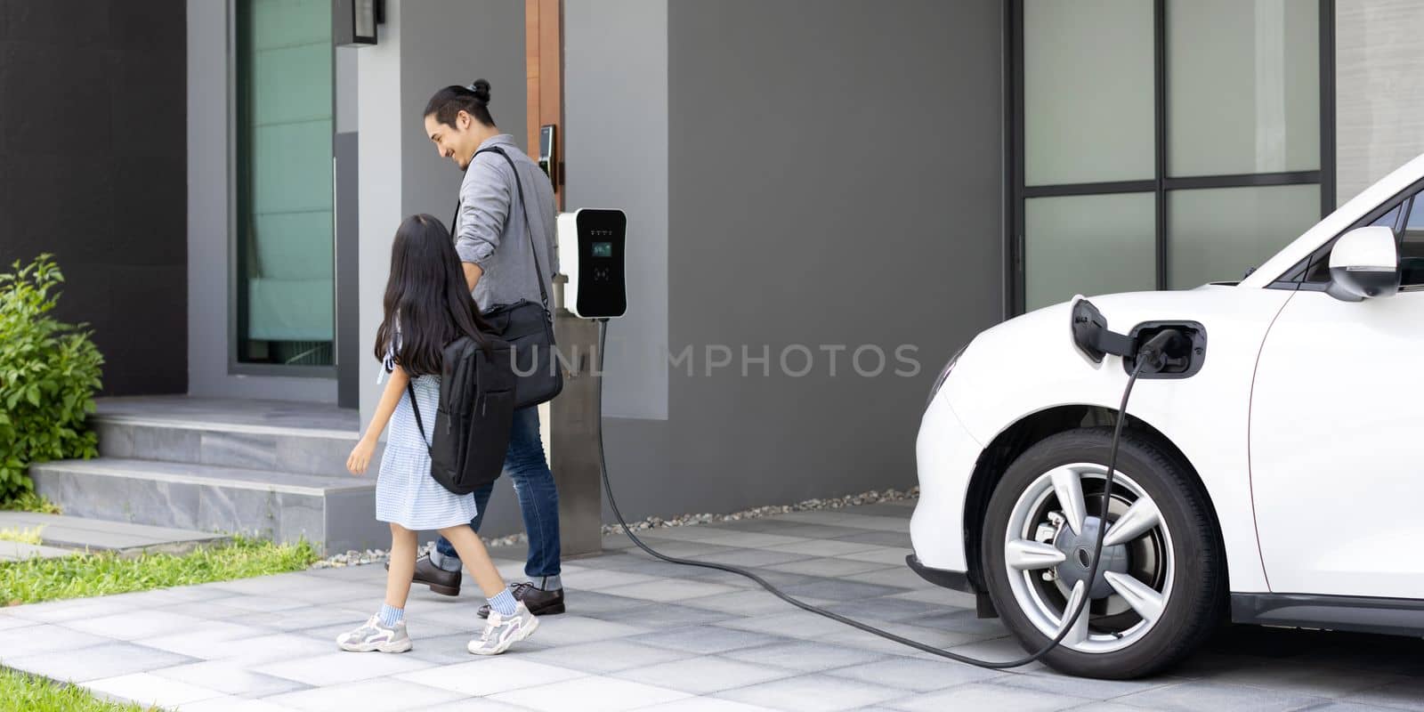 Progressive dad and daughter charging EV car from home charging station. by biancoblue