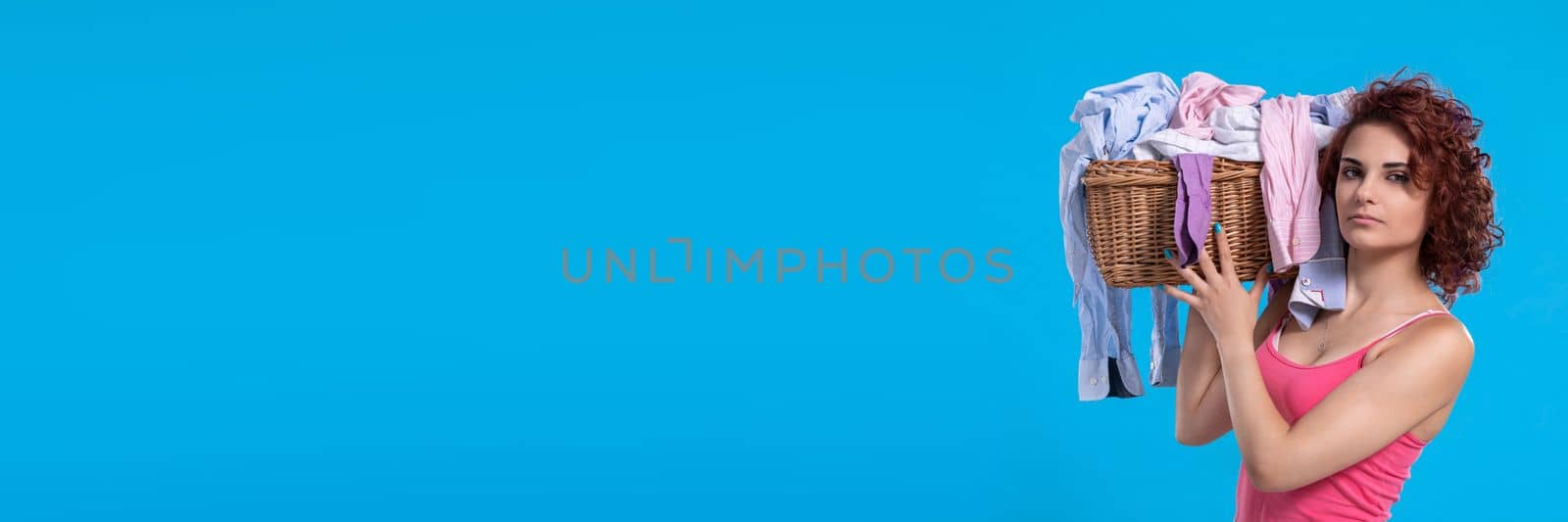 A strong woman holds a heavy wicker basket full of laundry clothes on her shoulder. An attractive girl with dark and curly hair. Panoramic view of girl with empty blue background.