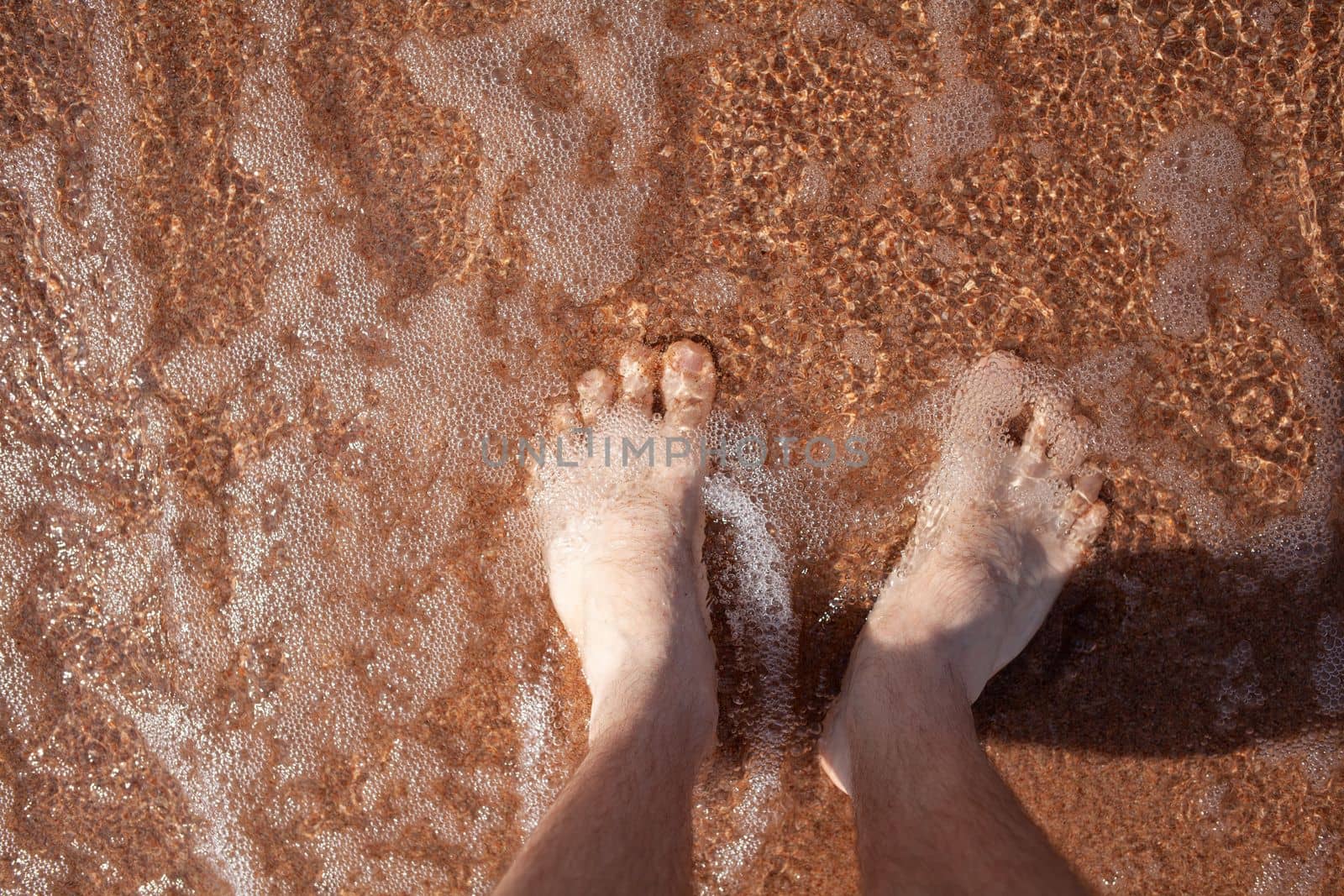 Top view of men's legs in the sea. A man is standing on the sand. An ocean wave washes the shore. Summer holidays.