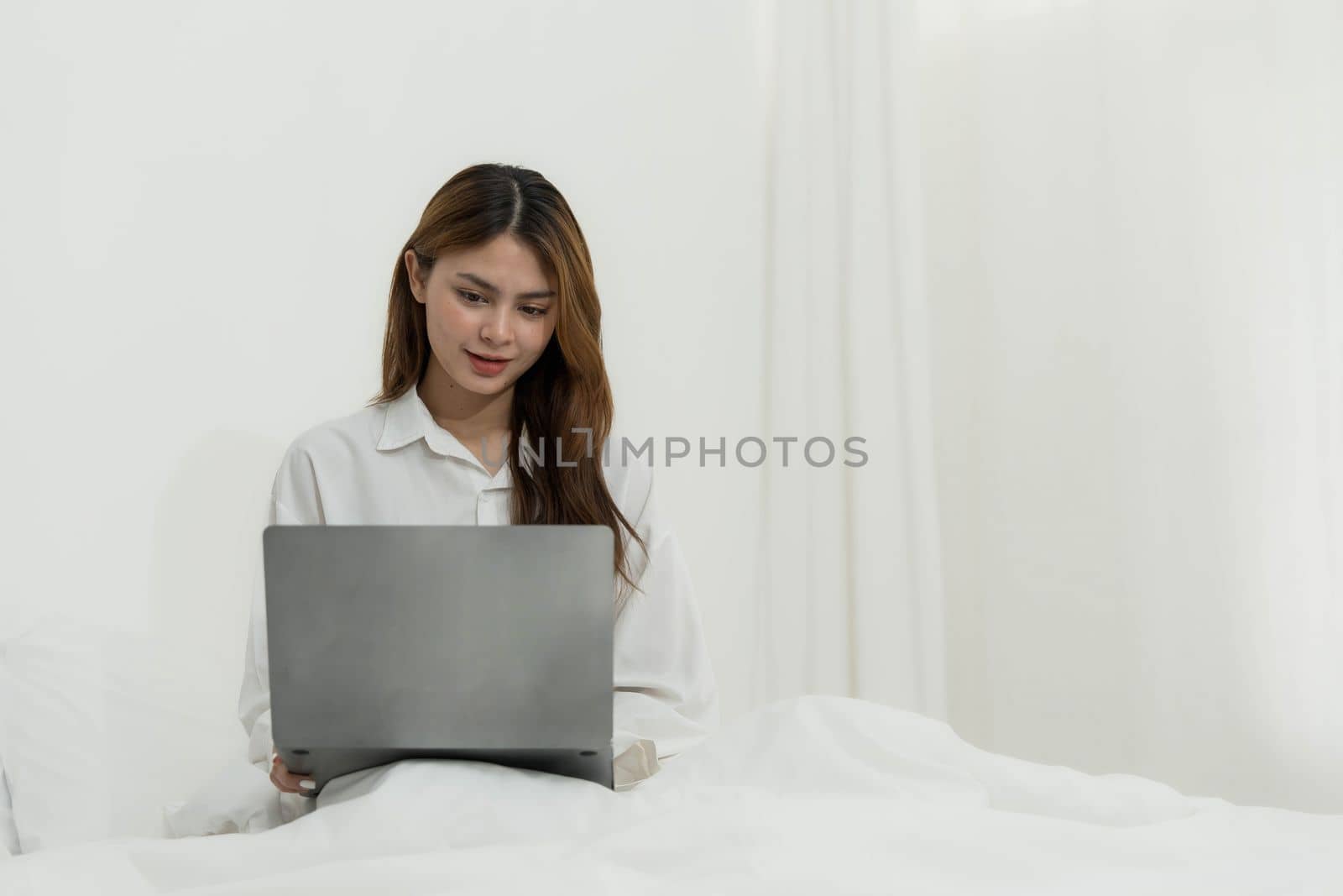 young asian woman using laptop when sitting on a bed. Smart living with communication technology for a better life. girl sitting on at white bedroom space with a clean interior design...