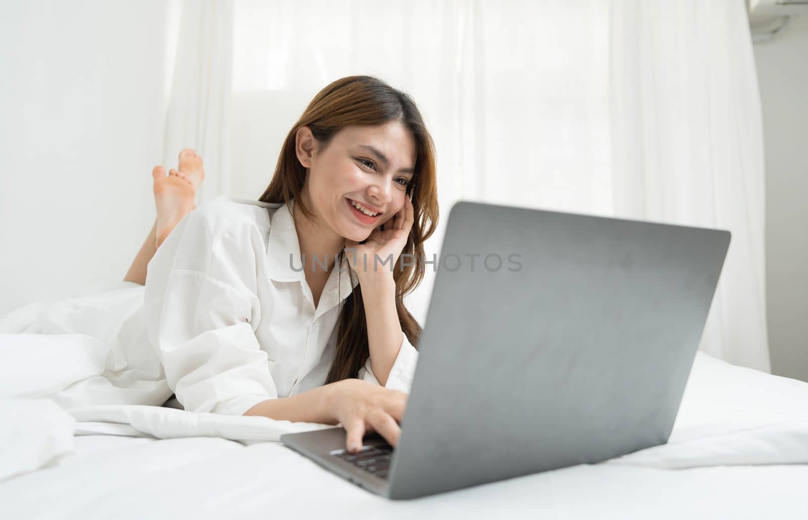 young asian woman using laptop when sitting on a bed. Smart living with communication technology for a better life. girl sitting on at white bedroom space with a clean interior design. by wichayada