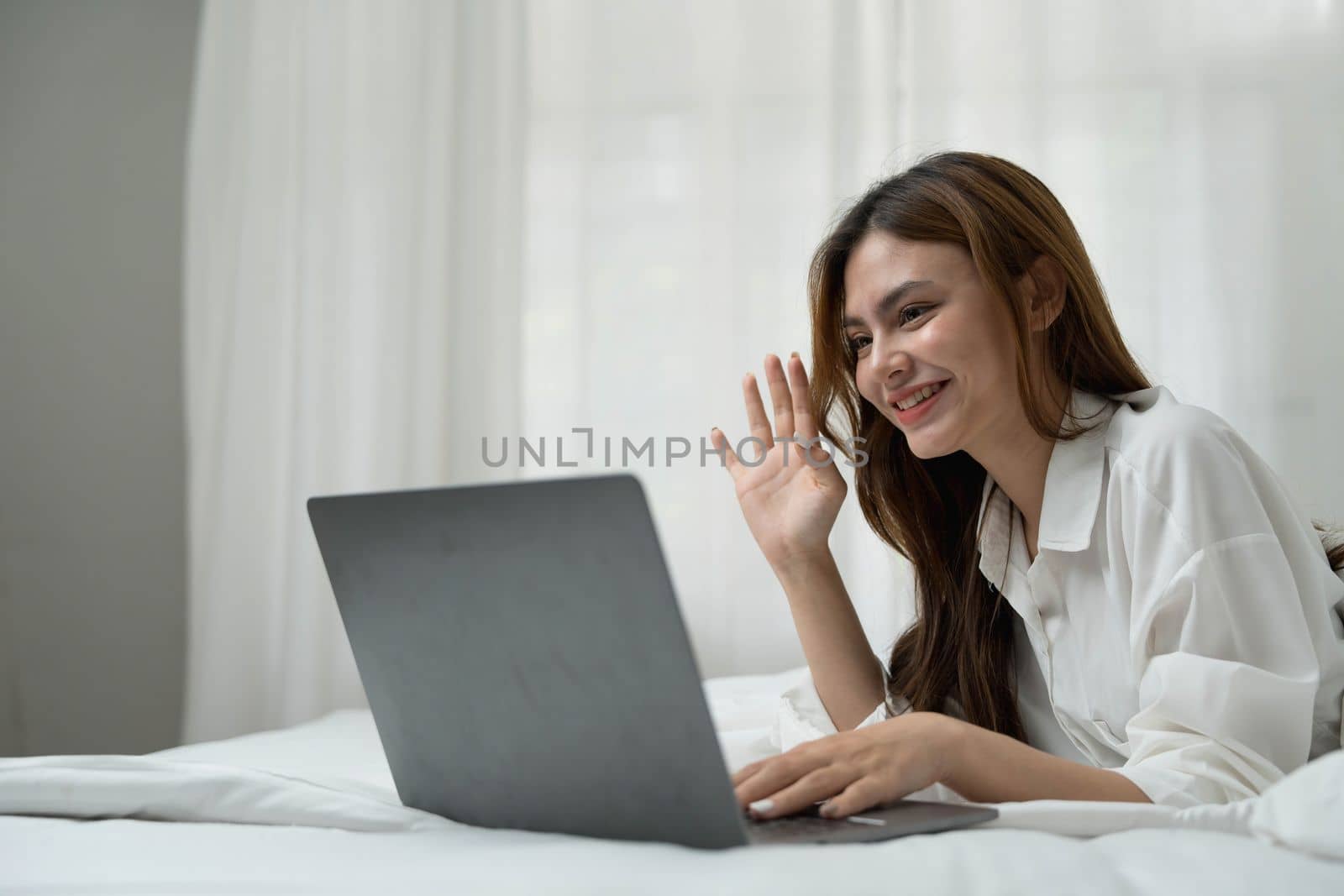 young asian woman using laptop when sitting on a bed. Smart living with communication technology for a better life. girl sitting on at white bedroom space with a clean interior design. by wichayada