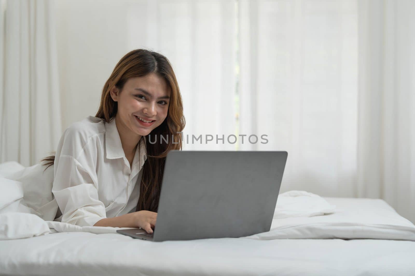 young asian woman using laptop when sitting on a bed. Smart living with communication technology for a better life. girl sitting on at white bedroom space with a clean interior design...