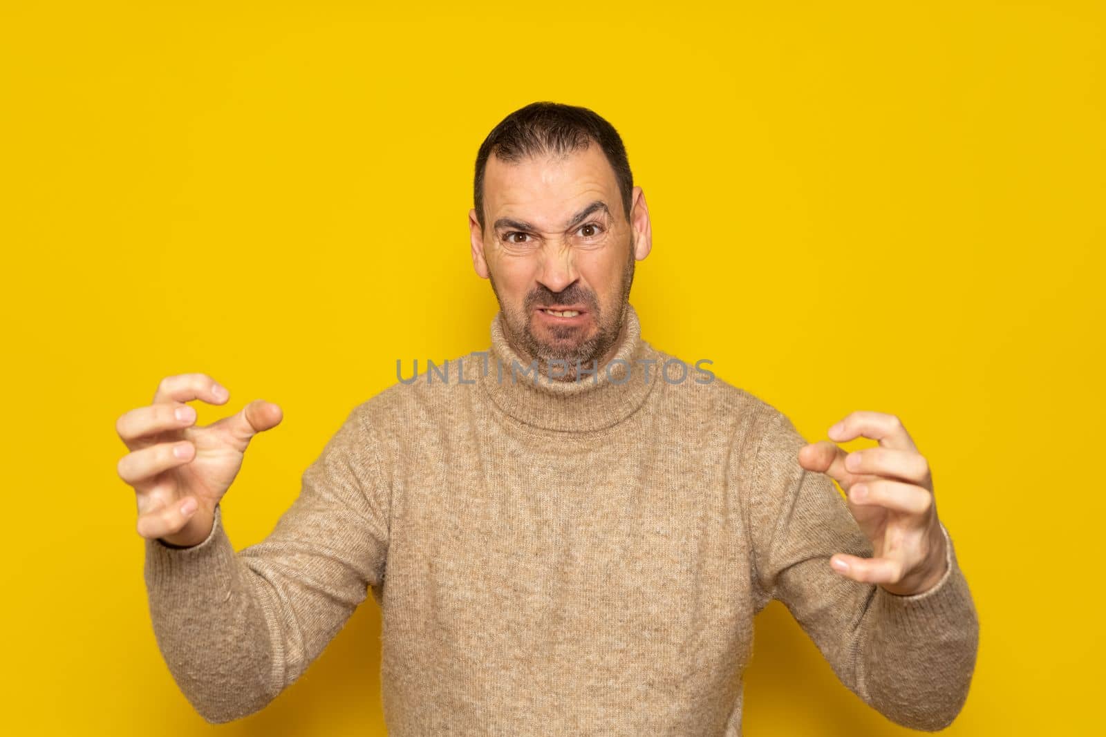 Hispanic man with beard over isolated yellow background in aggressive and furious attitude with raised hands in the shape of claws. Rage and irreverence concept
