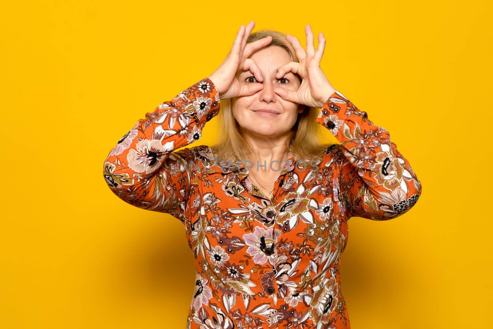 Portrait of astonished caucasian blonde woman spying, seeing amazing shocking event, looking through fingers imitating binoculars, wearing patterned dress. Isolated on yellow background.