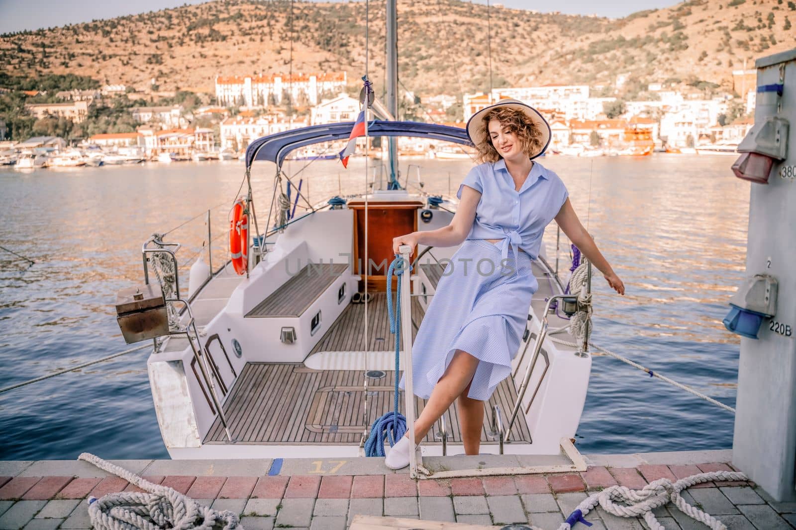 Woman yacht sea. A young happy woman in a blue dress and hat stands near the seaport with luxury yachts. Travel and vacation concept.