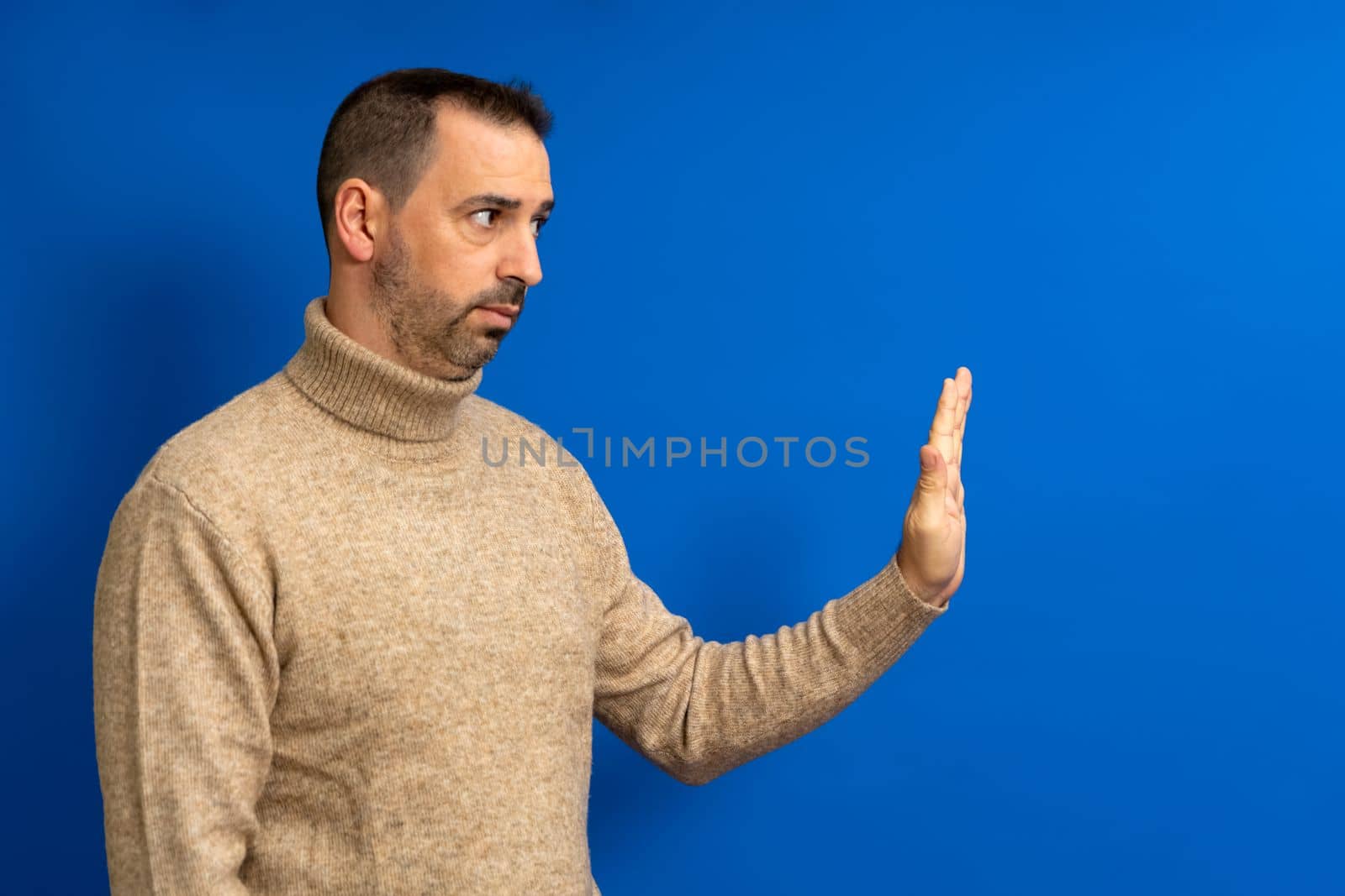 Strict bearded hispanic man showing stop sign, standing with frowning and bossy expression, meaning caution to avoid danger or mistake. Isolated on blue background.