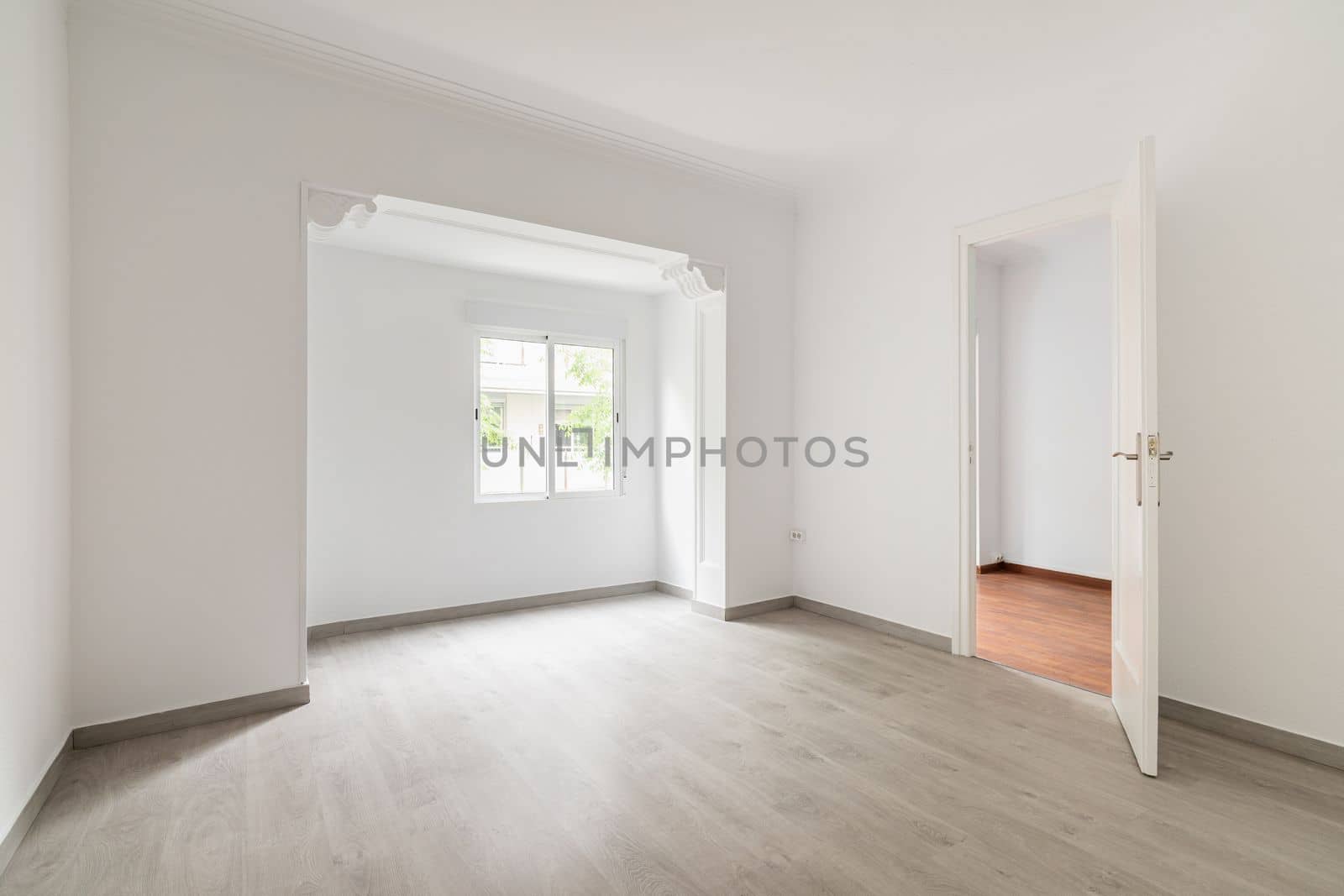 An empty spacious room, sunlight penetrates through the window during daylight hours. On floor there is wooden parquet in gray color. In open doorway is corridor to the rest of the apartment