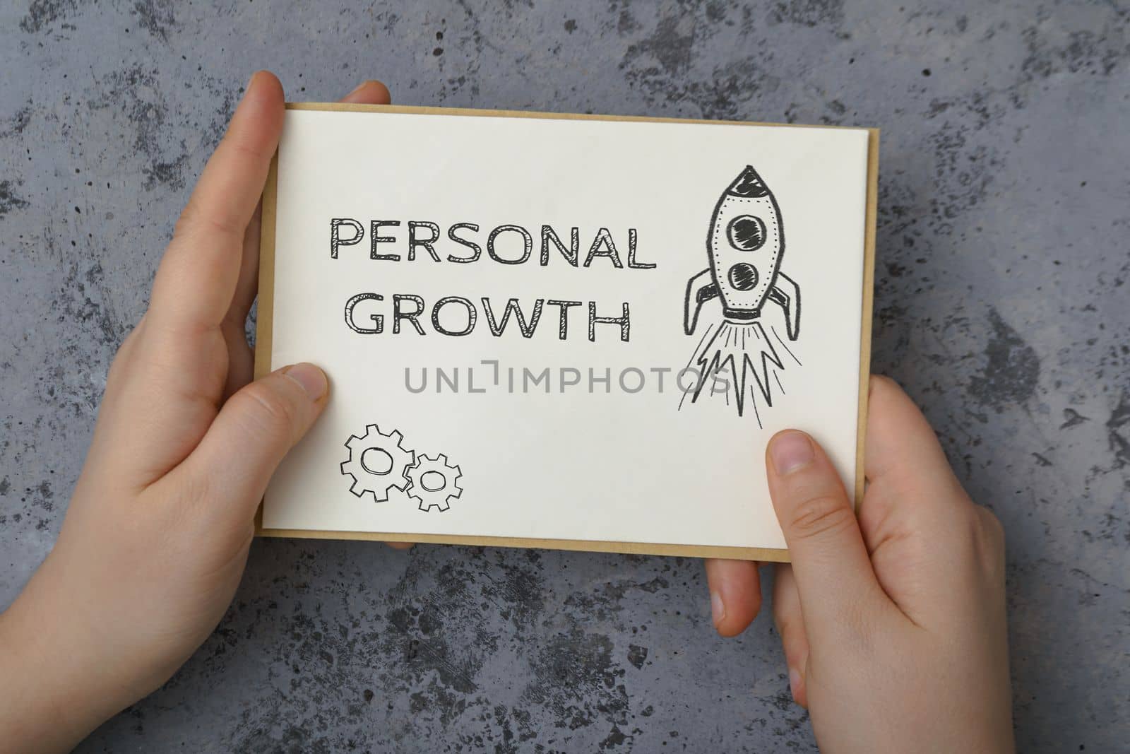 Person hands hold white card with phrase Personal Growth and drawn rocket against concrete background. Human resources and self-motivation in business