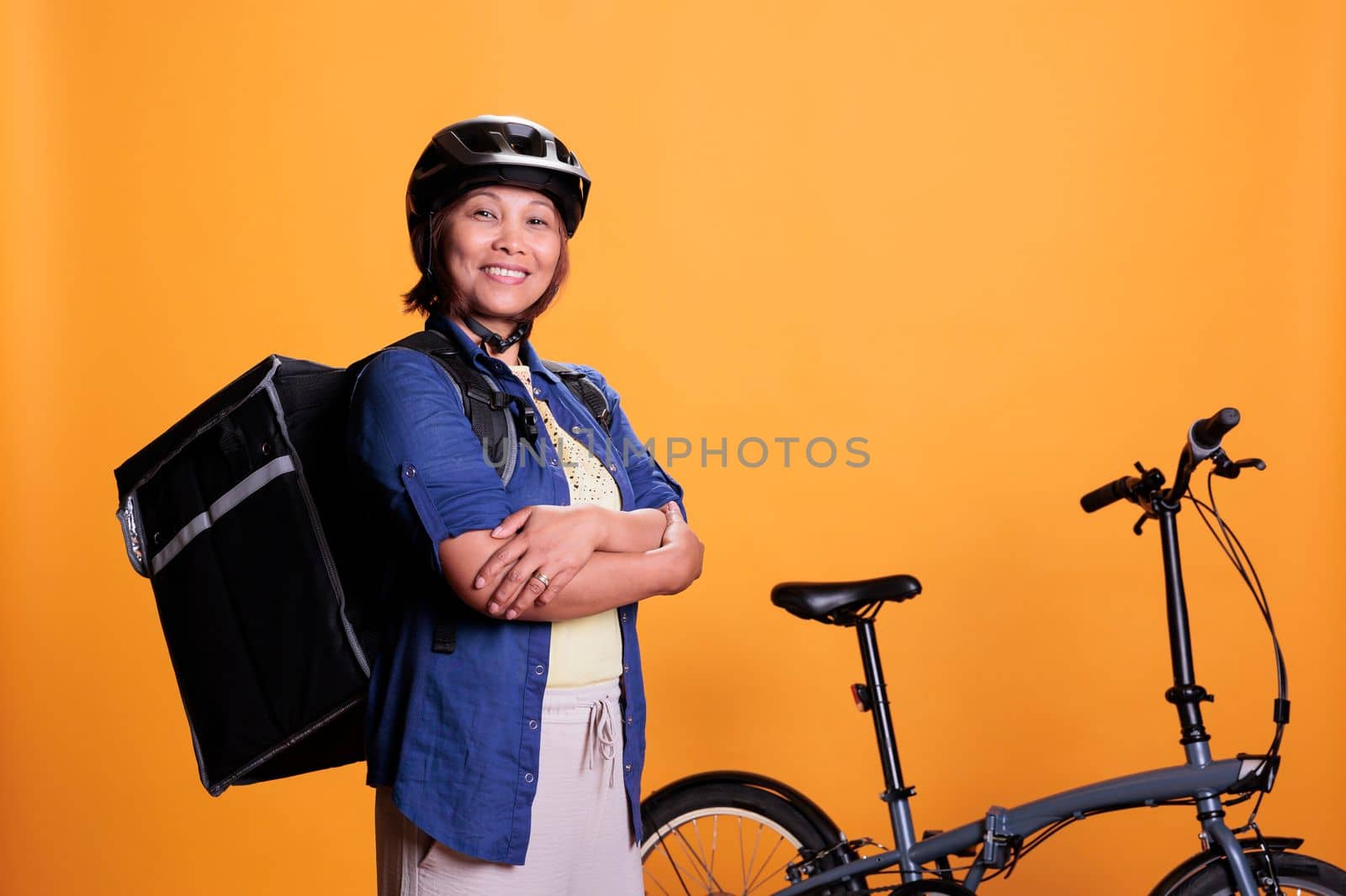 Deliver person carrying food delivery backpack standing beside bike in studio with yellow background. Restaurant worker delivering pizza for lunch. Food transportation service and takeaway concept