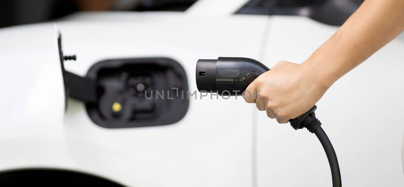 Focus hand holding EV charger plug with blurred background of progressive electric vehicle and socket parking in home garage with electric charging station powered by clean and sustainable energy.