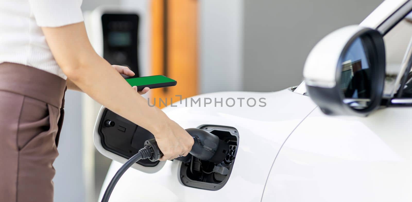 Closeup progressive woman install cable plug to her electric car with home charging station. Concept of the use of electric vehicles in a progressive lifestyle contributes to clean environment.