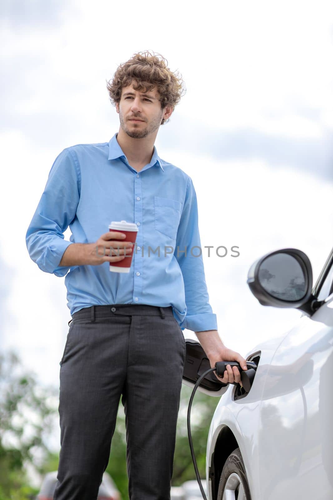 Progressive eco-friendly concept of parking EV car at public electric-powered charging station in city with blur background of businessman leaning on recharging-electric vehicle with coffee.