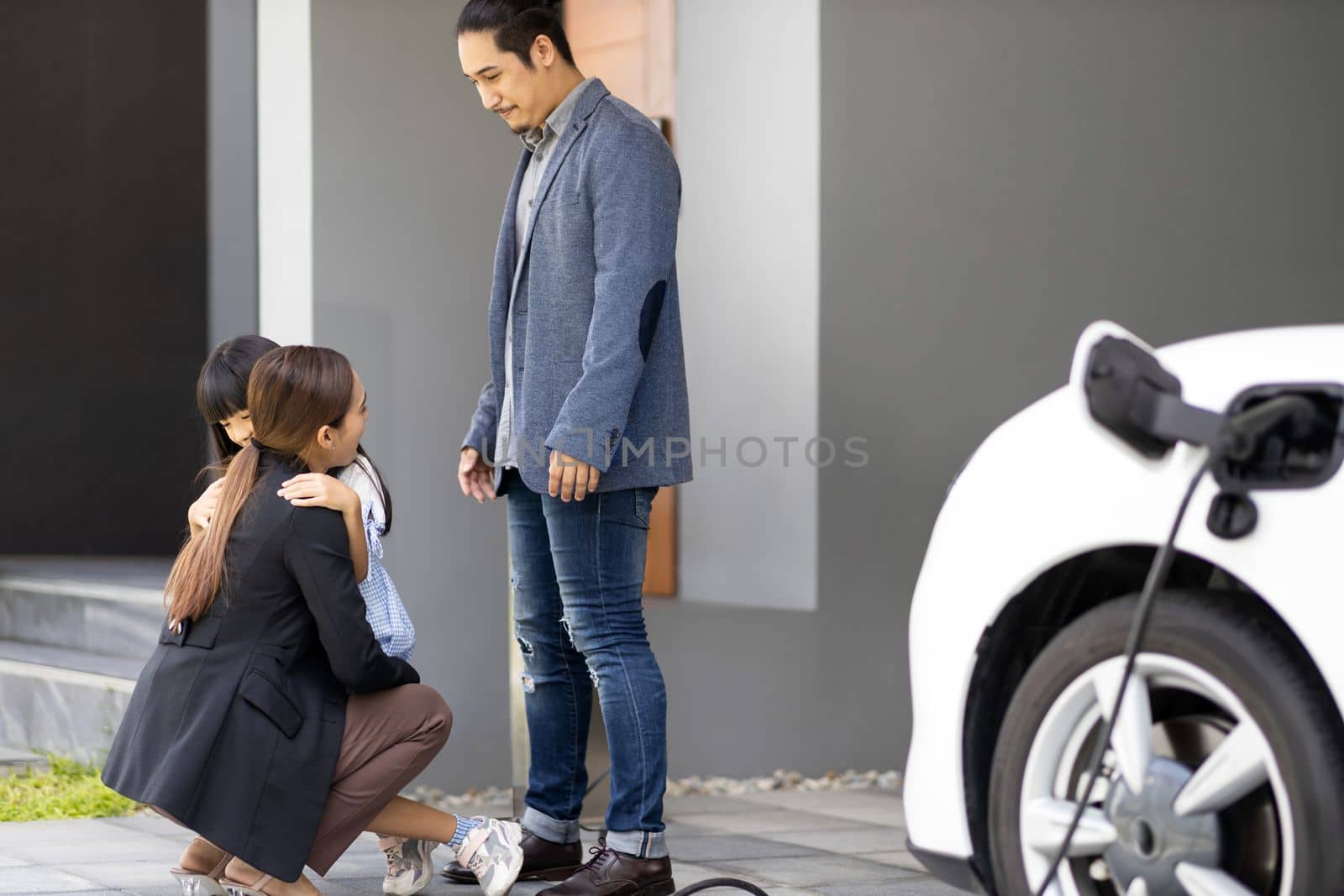 Progressive young parents and daughter with electric vehicle and home charging station. Green and clean energy from electric vehicles for healthy environment. Eco power from renewable source at home.