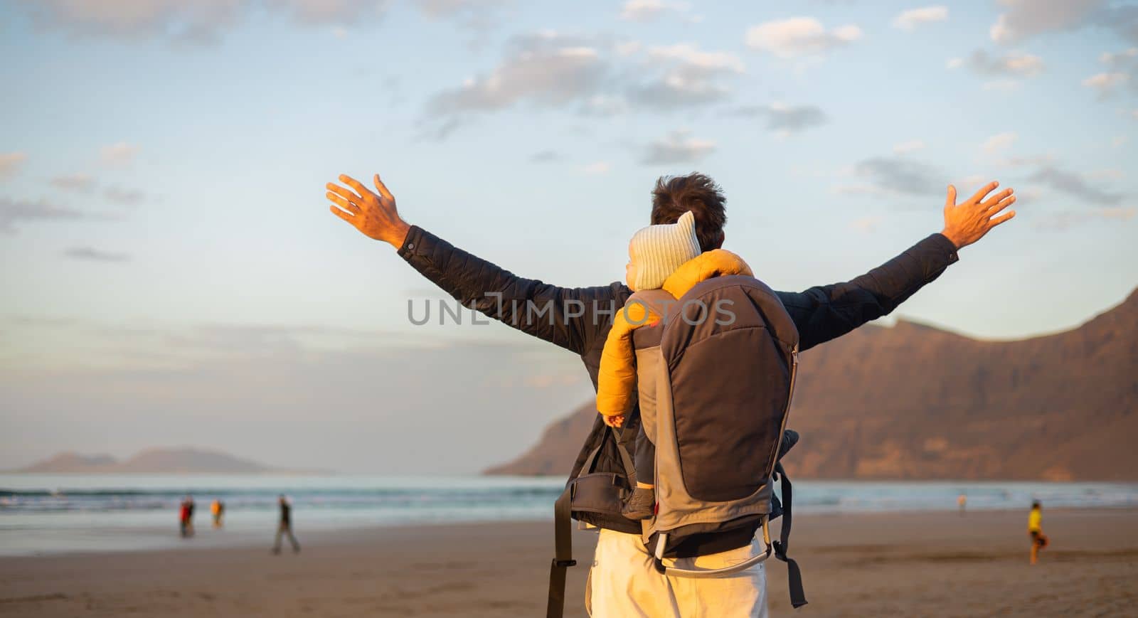 Young father rising hands to the sky while enjoying pure nature carrying his infant baby boy son in backpack on windy sandy beach of Famara, Lanzarote island, Spain at sunset. Family travel concept. by kasto